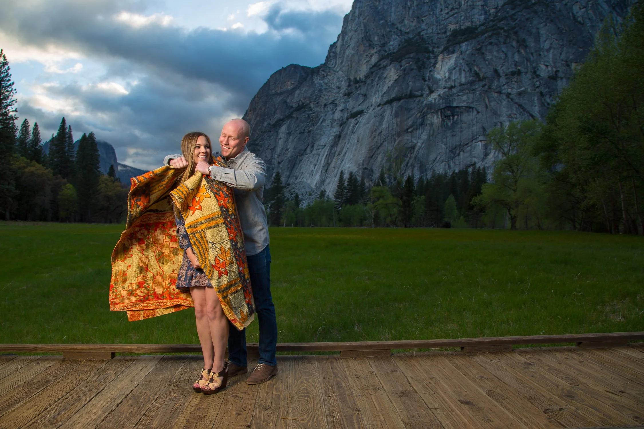 A couple stands on a wooden platform and embraces each other, wrapped in a colorful, patterned blanket, with a scenic mountain landscape, trees, and cloudy sky in the background.