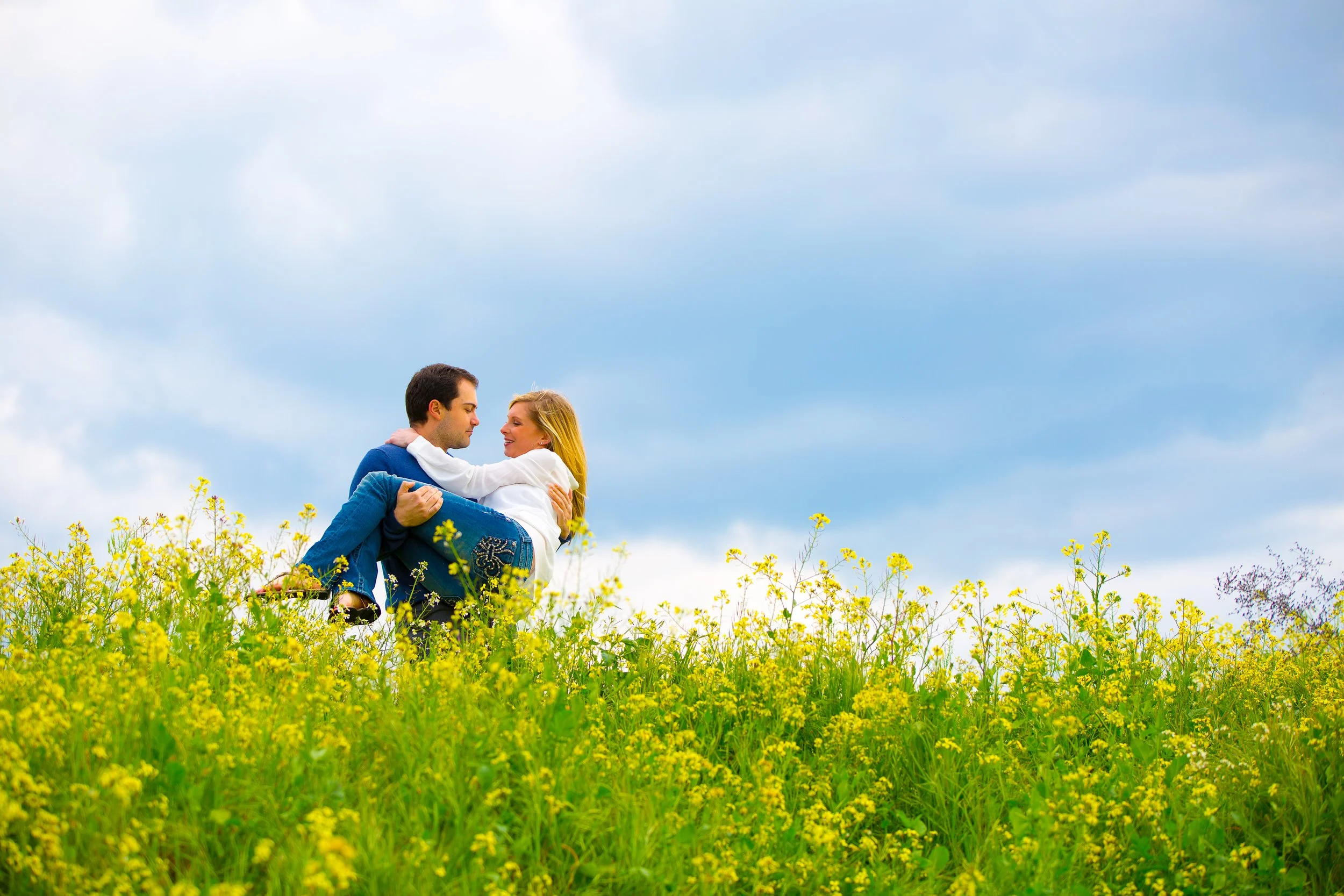 A couple sitting in a yellow flower field, smiling and looking at each other against a blue sky with clouds.