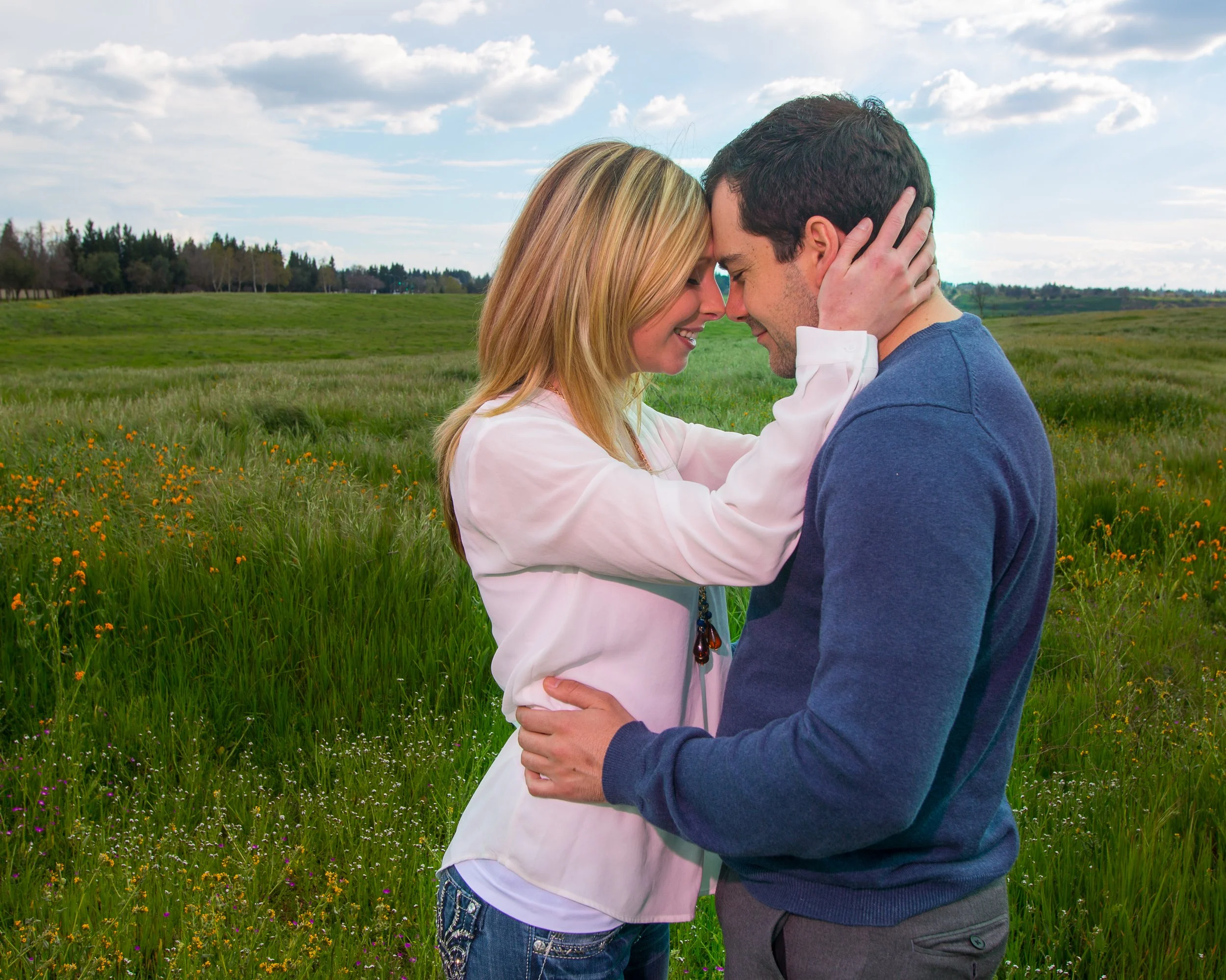 A couple standing in a grassy field with their foreheads touching and smiling, holding each other lovingly.