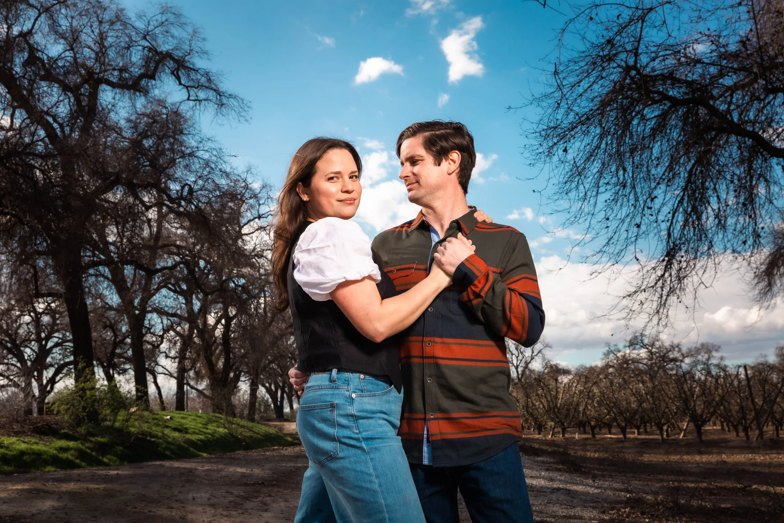 A young couple dancing outdoors in a park during daytime, with leafless trees and a partly cloudy sky in the background.