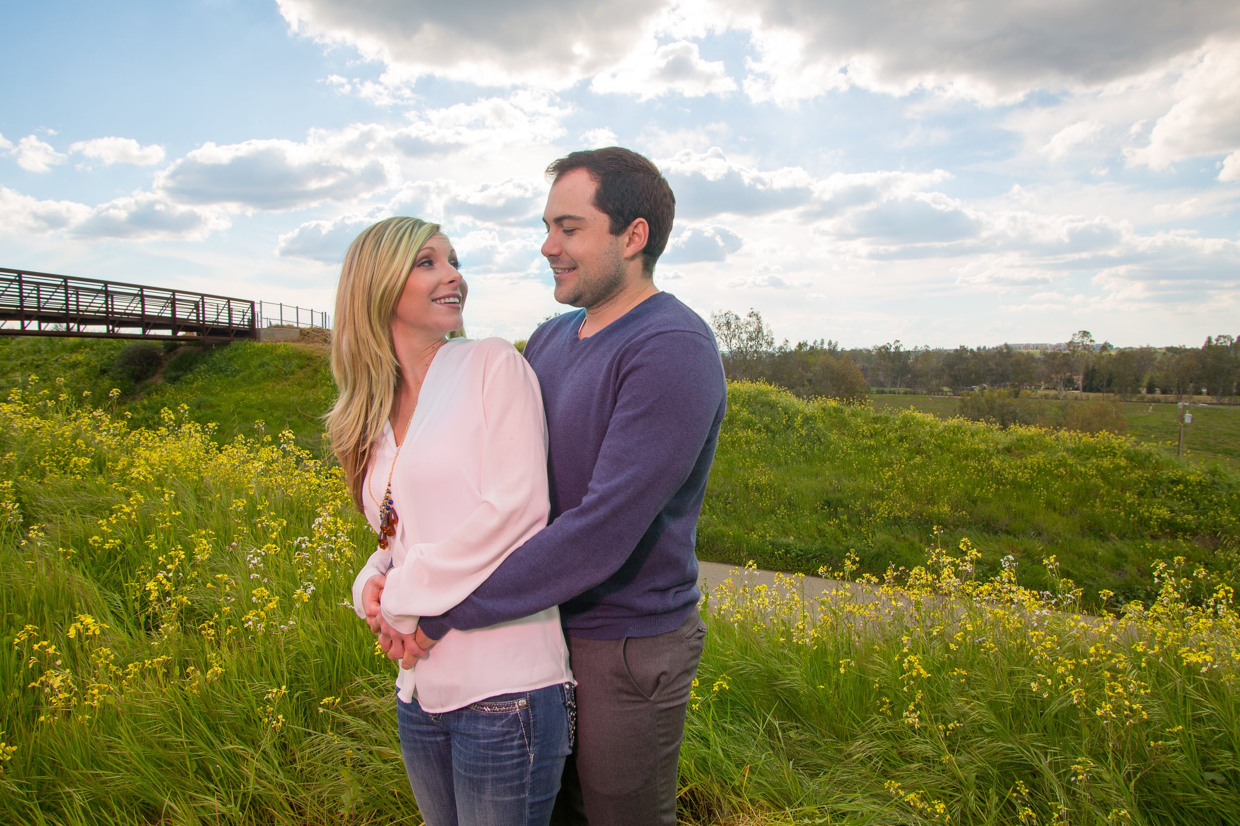 A couple standing in a field of yellow flowers, holding hands and looking at each other, with a bridge and cloudy sky in the background.