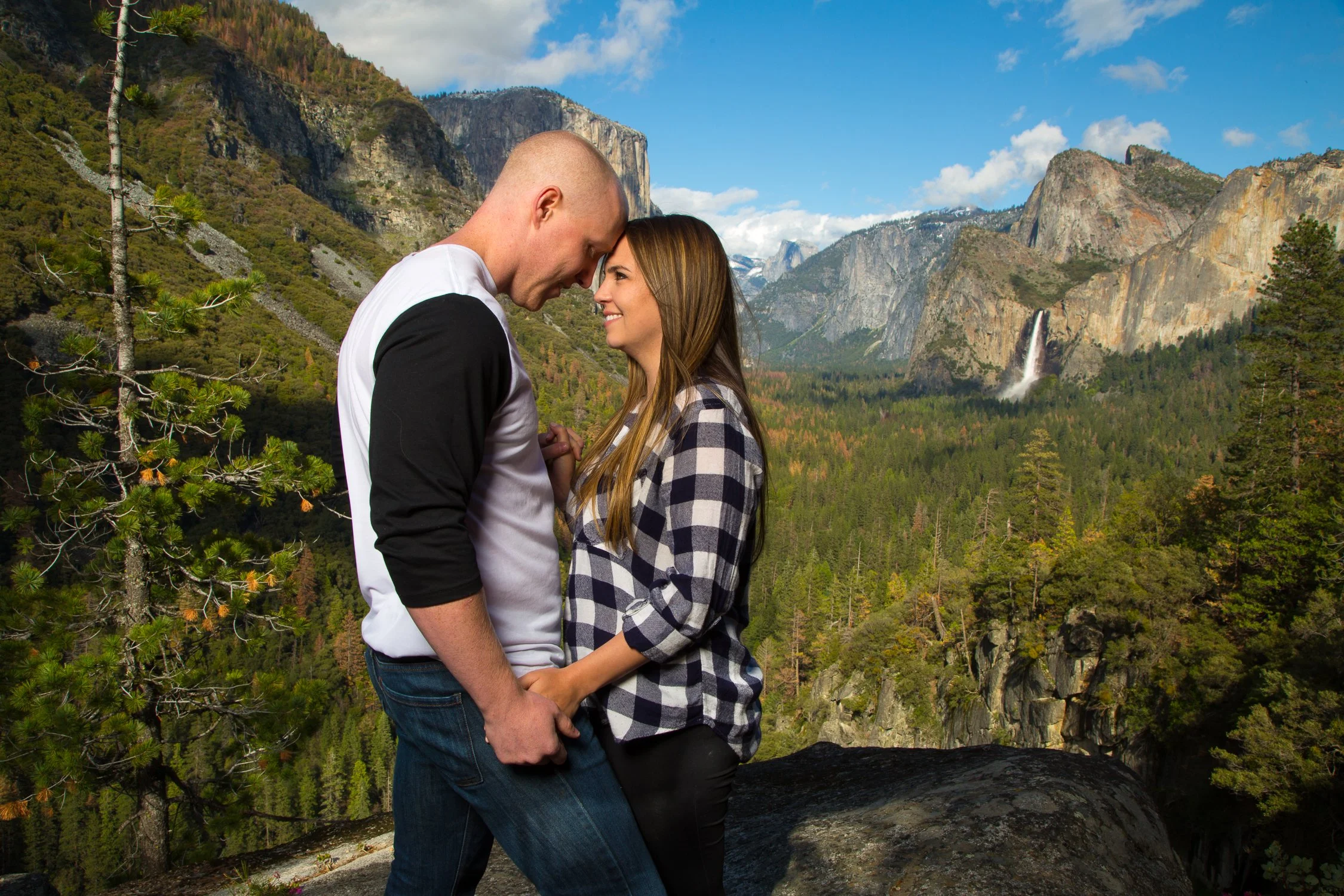 A couple standing close together outdoors with a mountain landscape, trees, and a waterfall in the background.