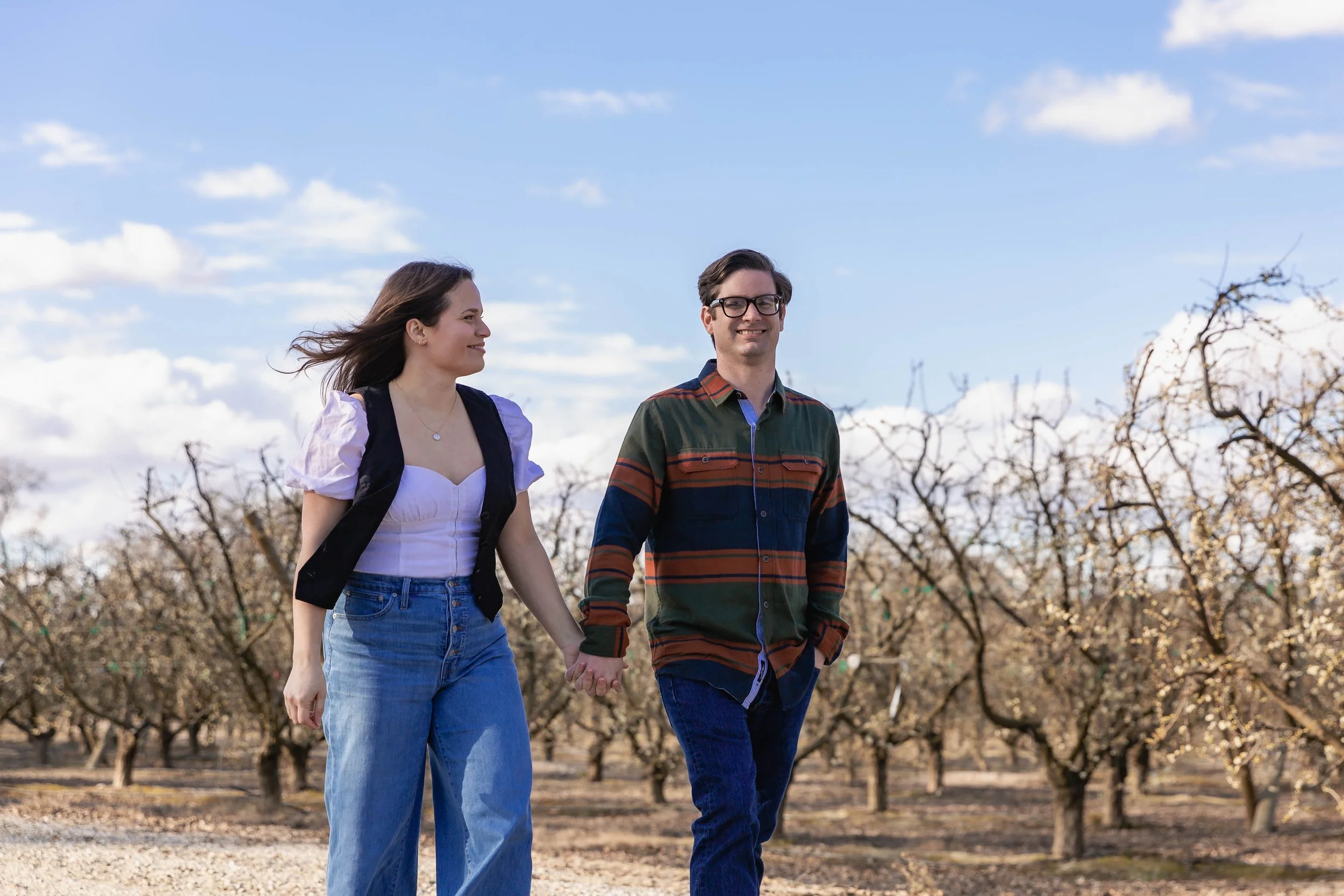 A couple holding hands and walking through an orchard with bare trees under a partly cloudy sky.