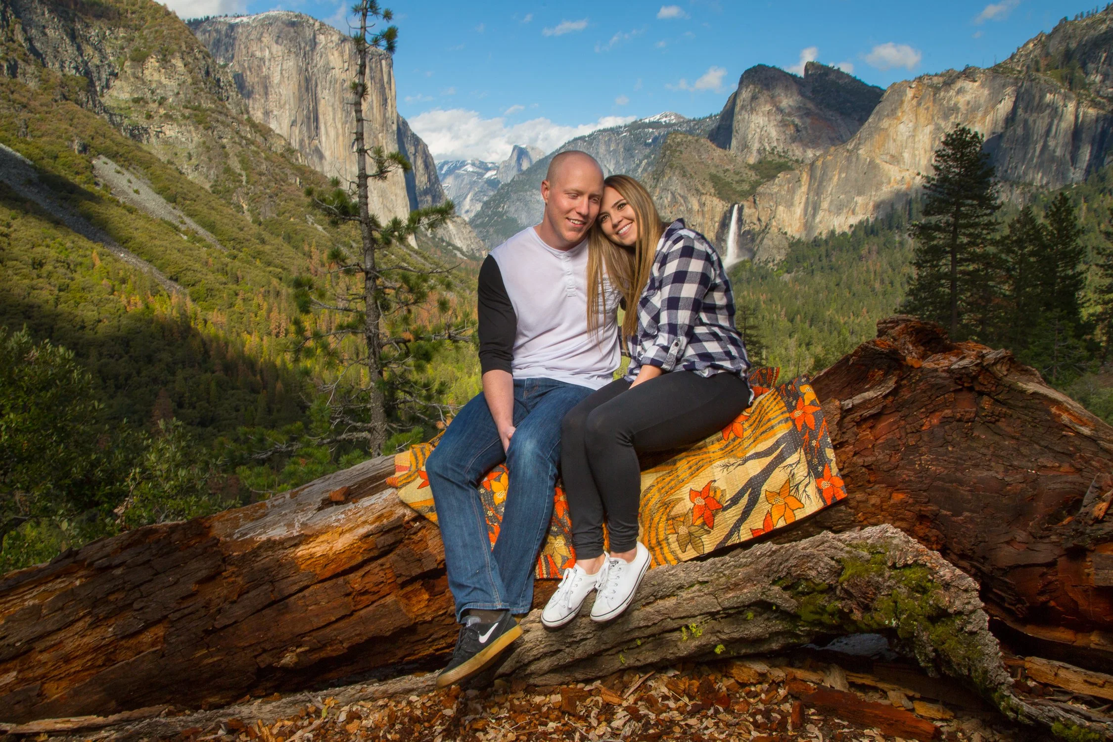 A young couple sitting on a large fallen tree trunk in a mountain landscape with trees, cliffs, and a waterfall in the background.