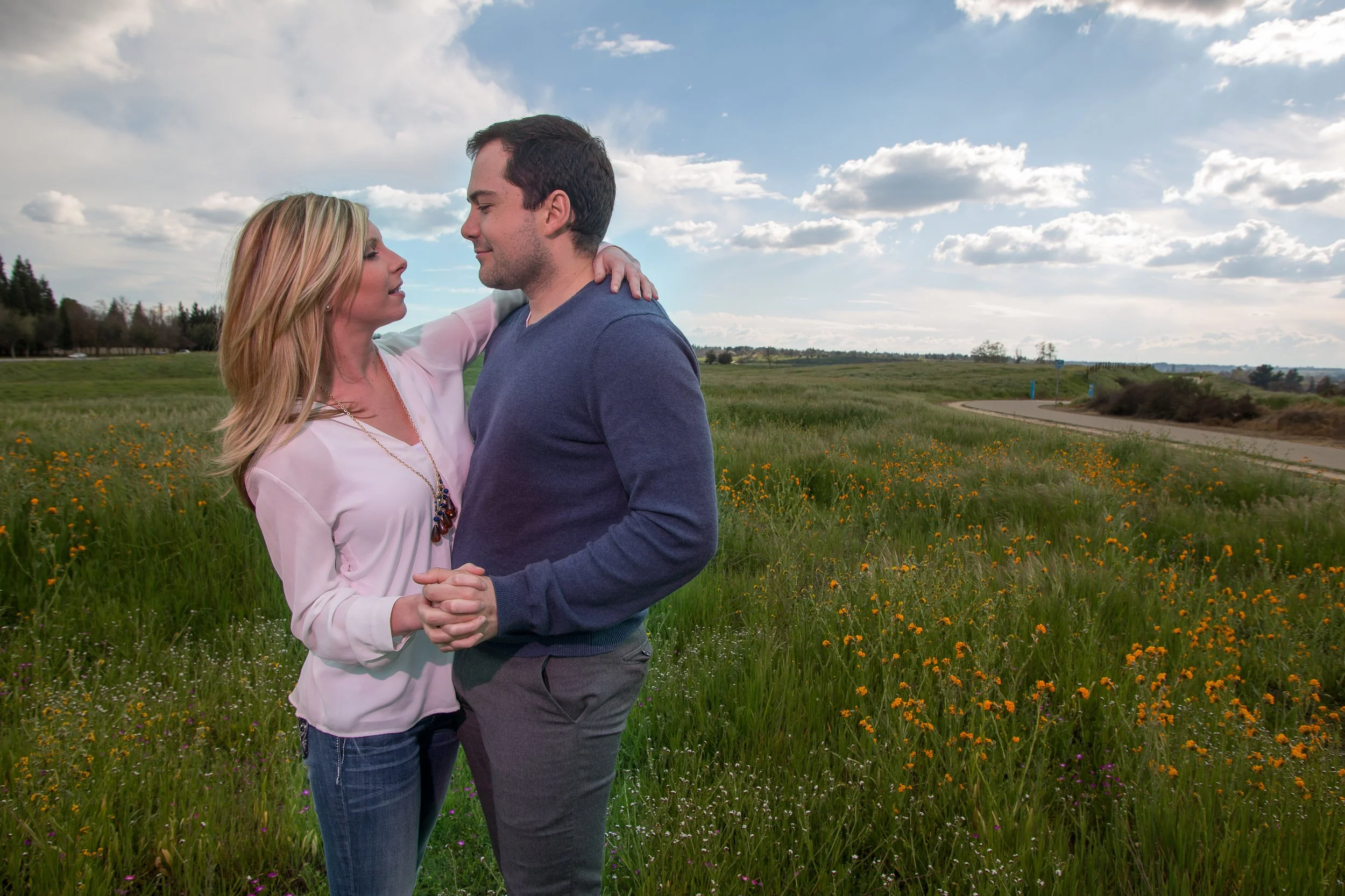 A couple dancing outdoors in a field with green grass, orange wildflowers, and a winding road under a partly cloudy sky.