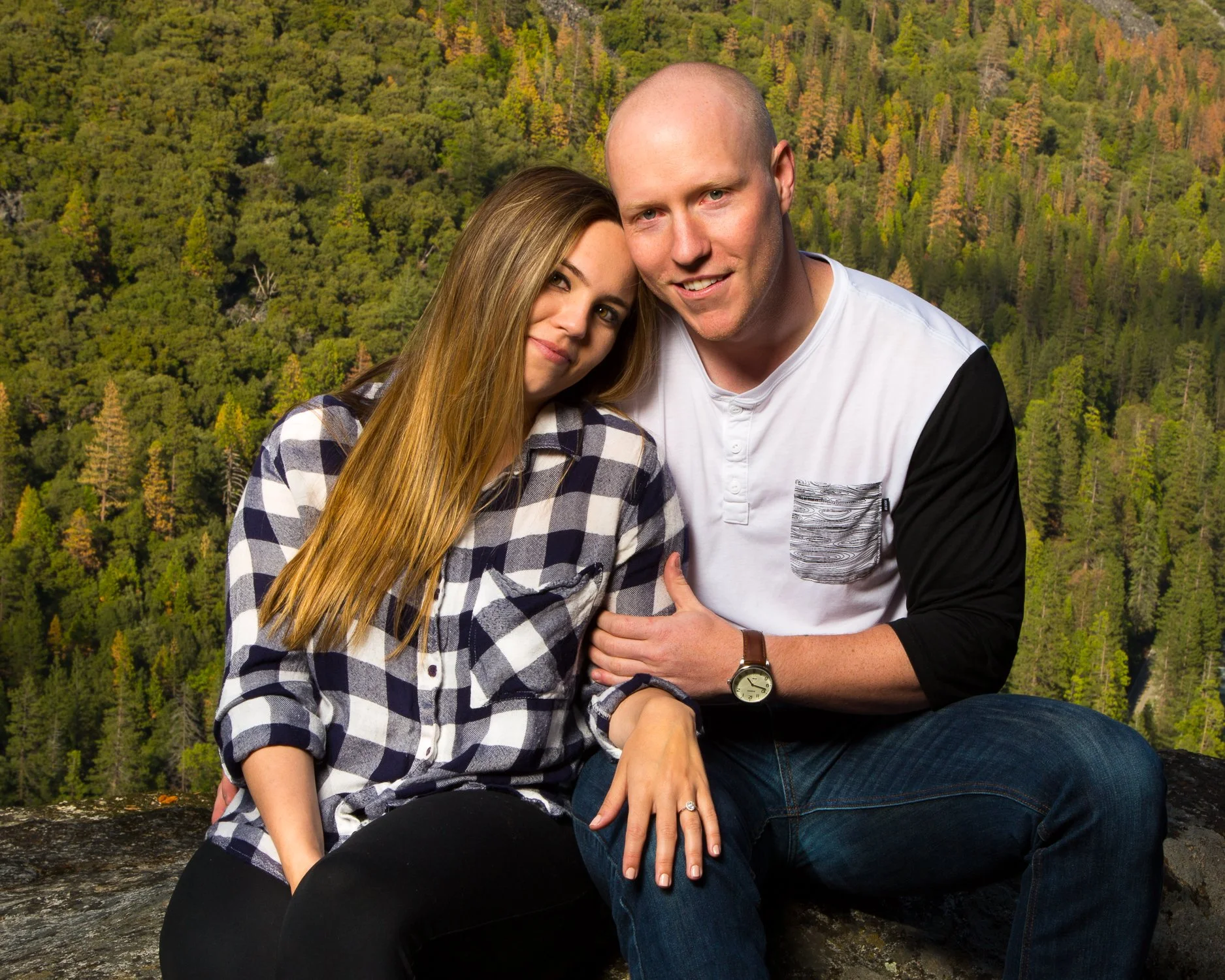 A young couple sitting outdoors on a rock, with a lush green forested mountain in the background. The woman has long blonde hair and is wearing a black and white checkered shirt. The man is bald, wearing a white and black long-sleeve shirt, and has a