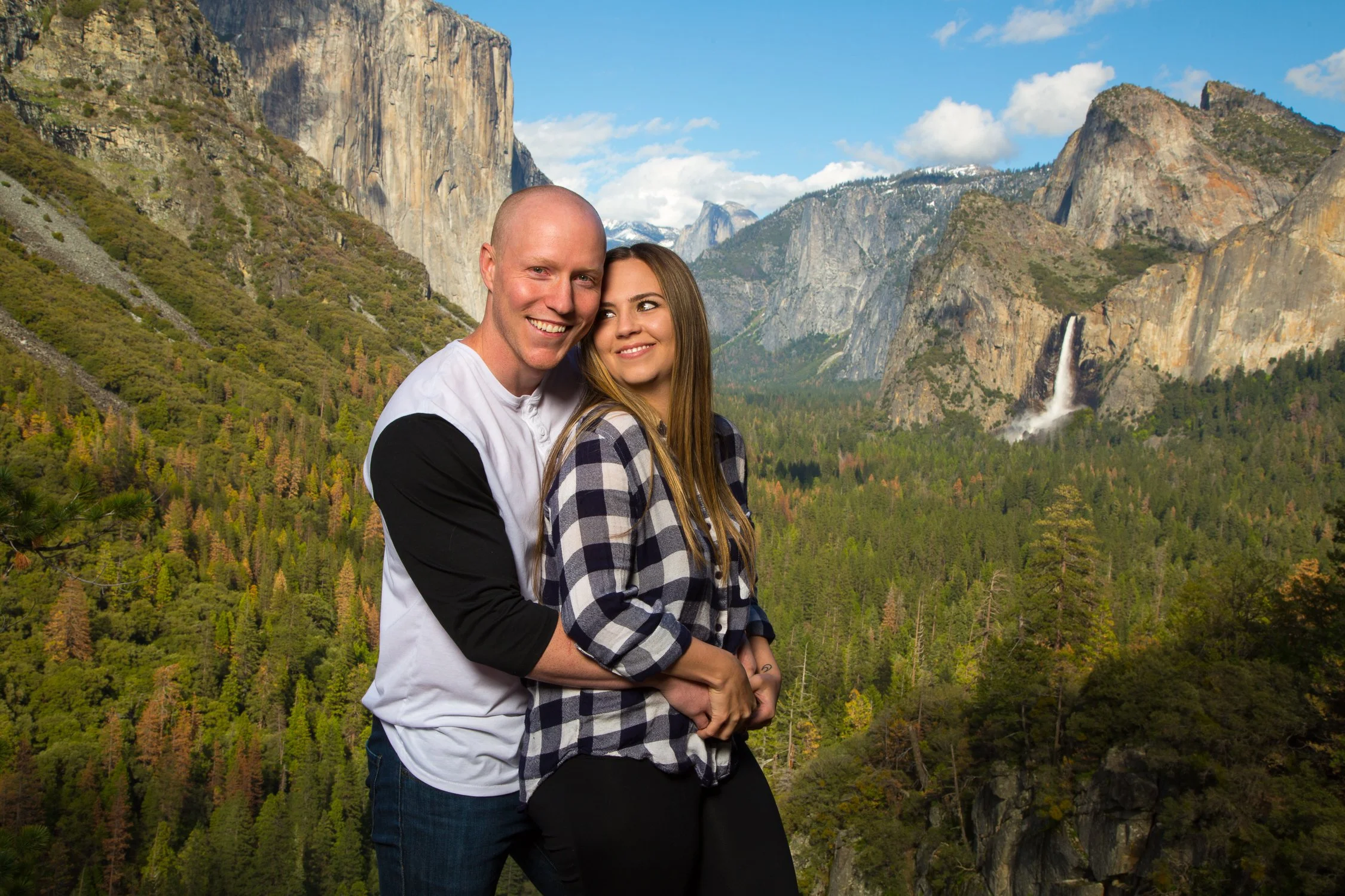 A couple standing together outdoors in front of a scenic landscape with mountains, a waterfall, and a forest under a partly cloudy sky.