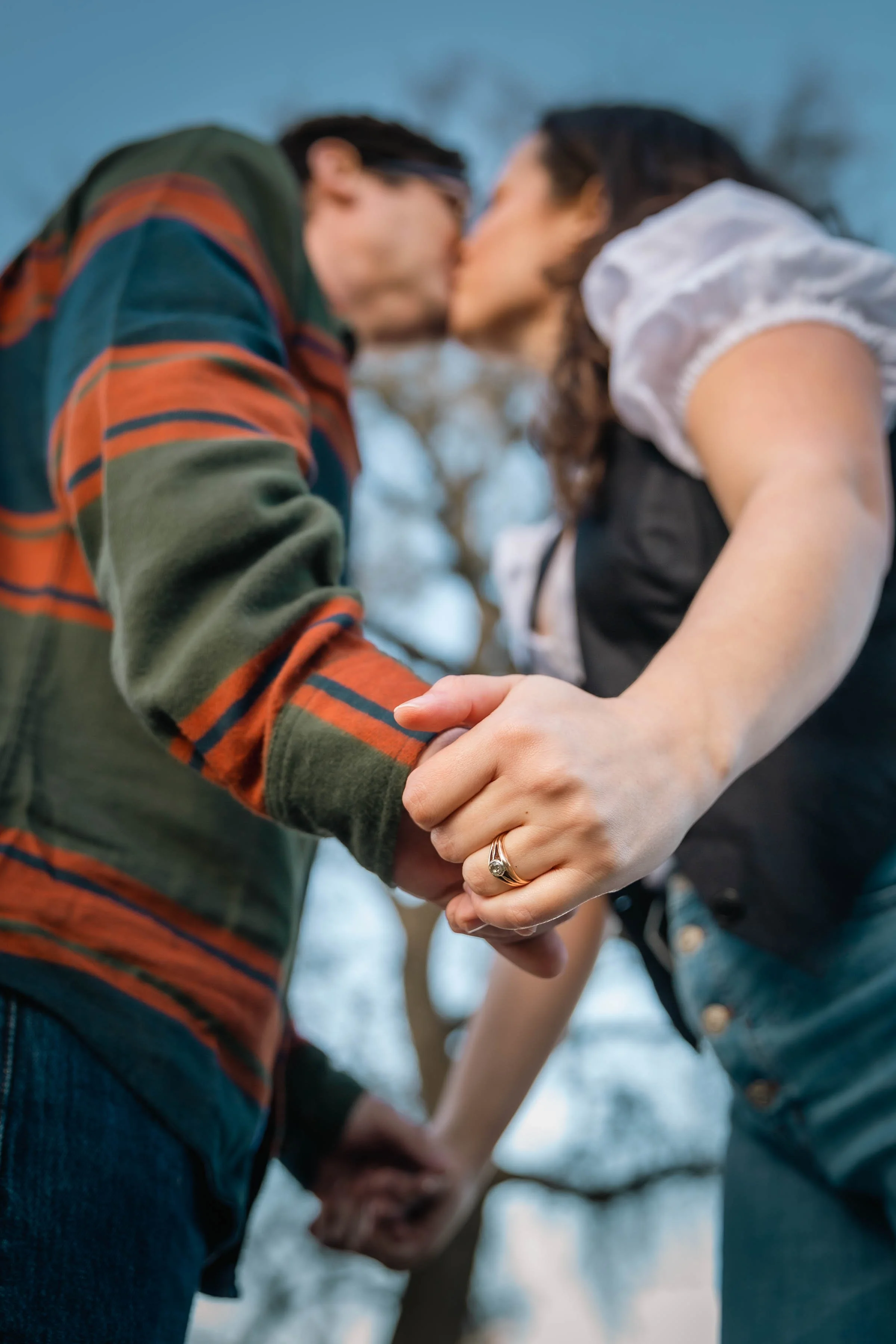 A couple holding hands and kissing outdoors, with focus on their hands and a wedding ring on the woman's finger.
