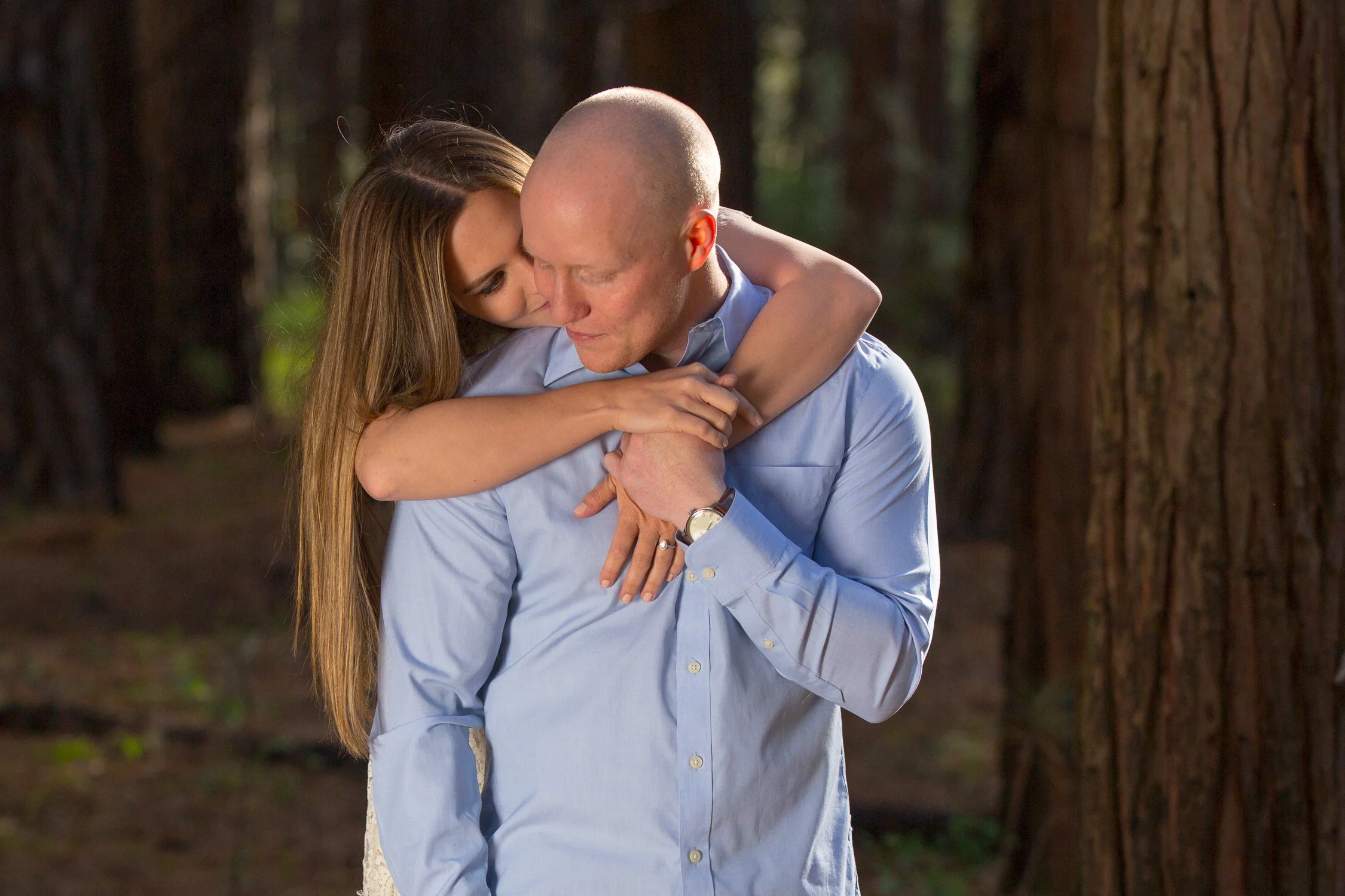 A young woman hugging a bald man in a forest, with large tree trunks visible in the background.