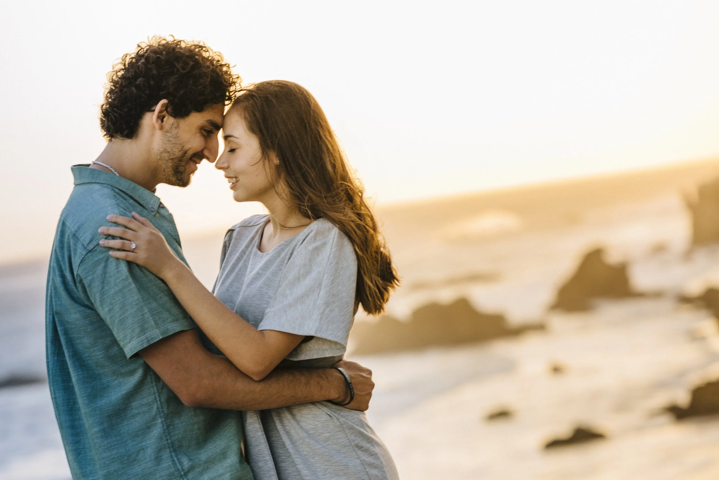 A young couple with light skin and dark hair embrace on a beach during sunset, smiling with foreheads touching.