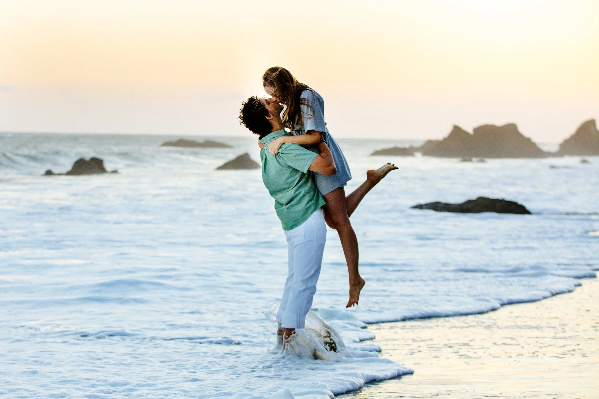 A couple on the beach, with the man lifting the woman, as the sun sets over the ocean.
