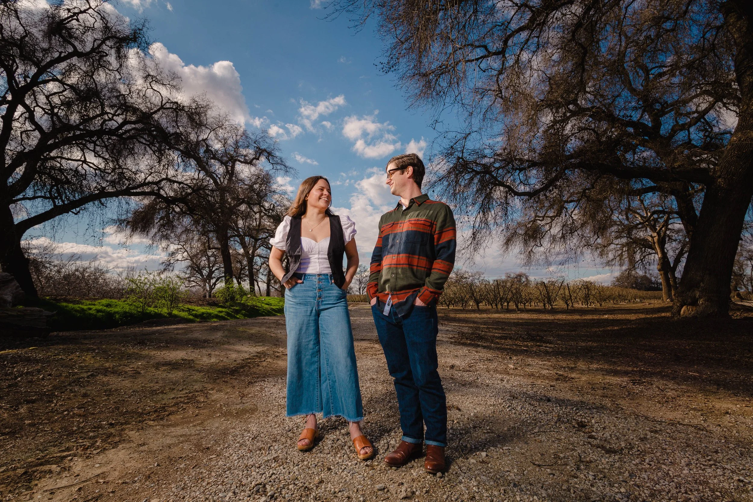 A woman and a man standing on a dirt path in a park with trees, smiling at each other under a partly cloudy sky.