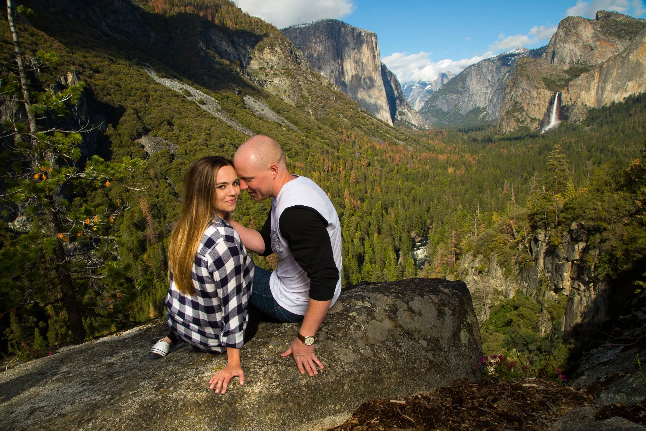 A couple sitting on a large rock overlooking a forested valley with tall cliffs and waterfalls in the distance.