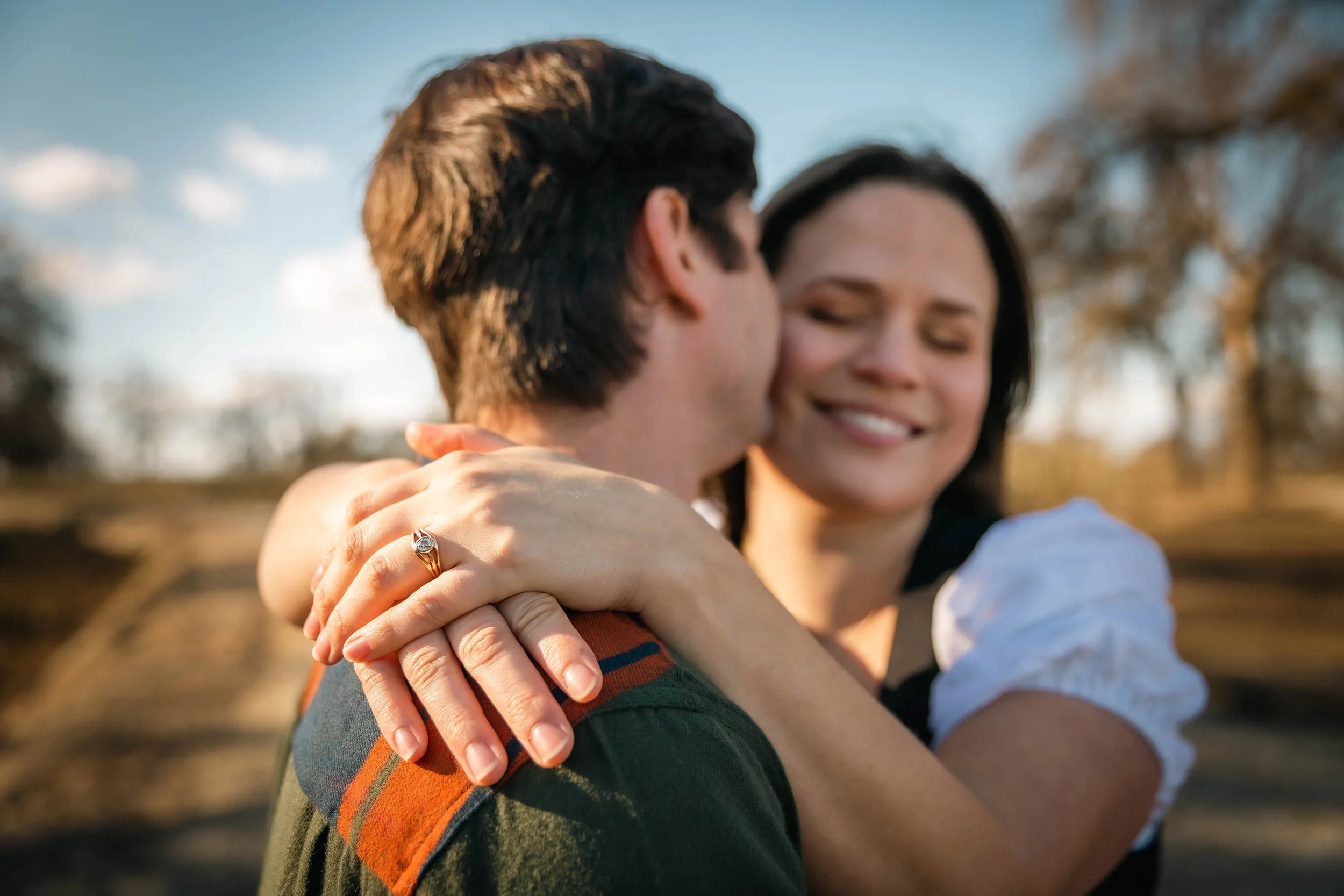 A couple hugging outdoors during sunset, with the woman smiling and wearing an engagement ring.