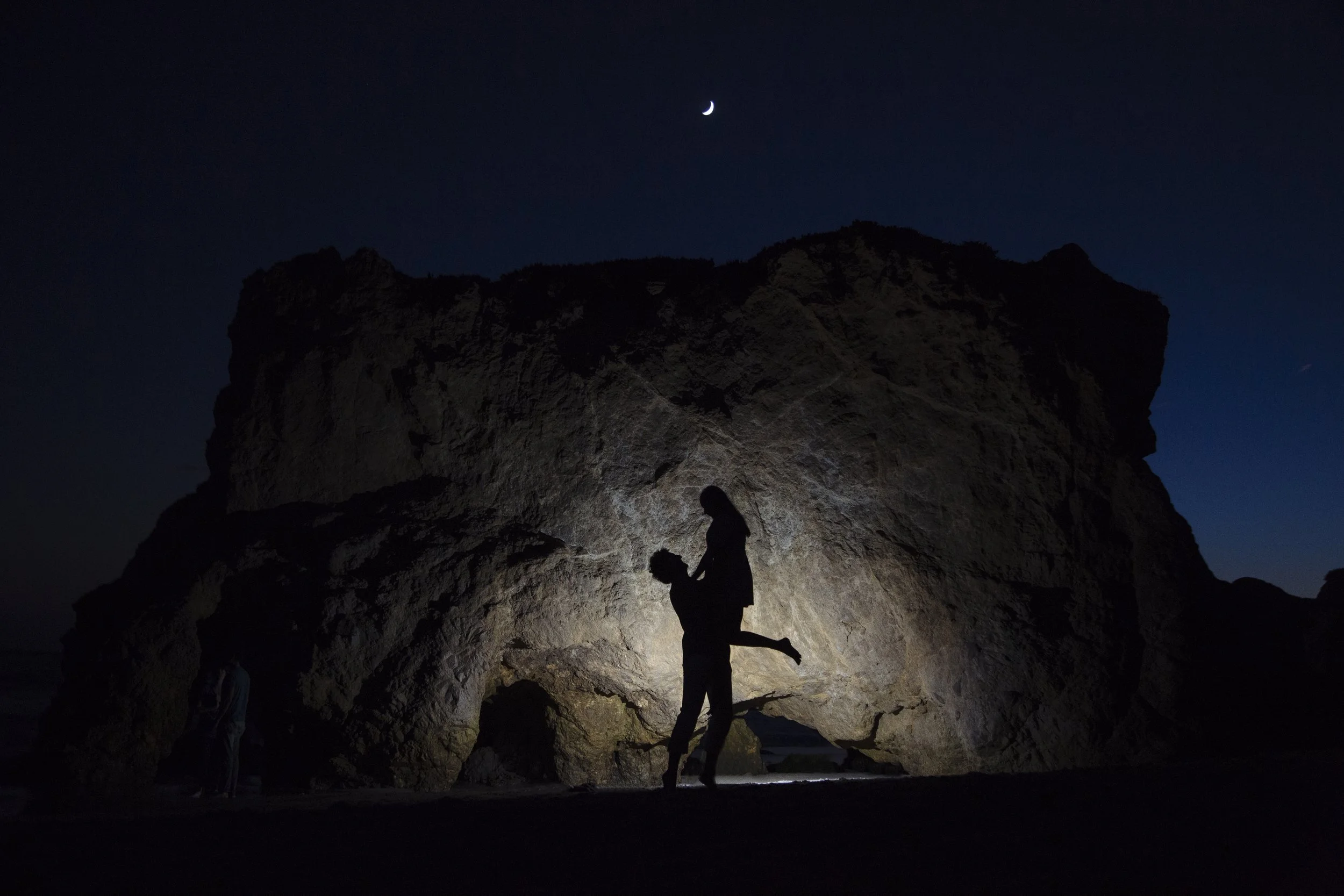 Silhouette of a person carrying another person on their back against a large cave and night sky with a crescent moon.
