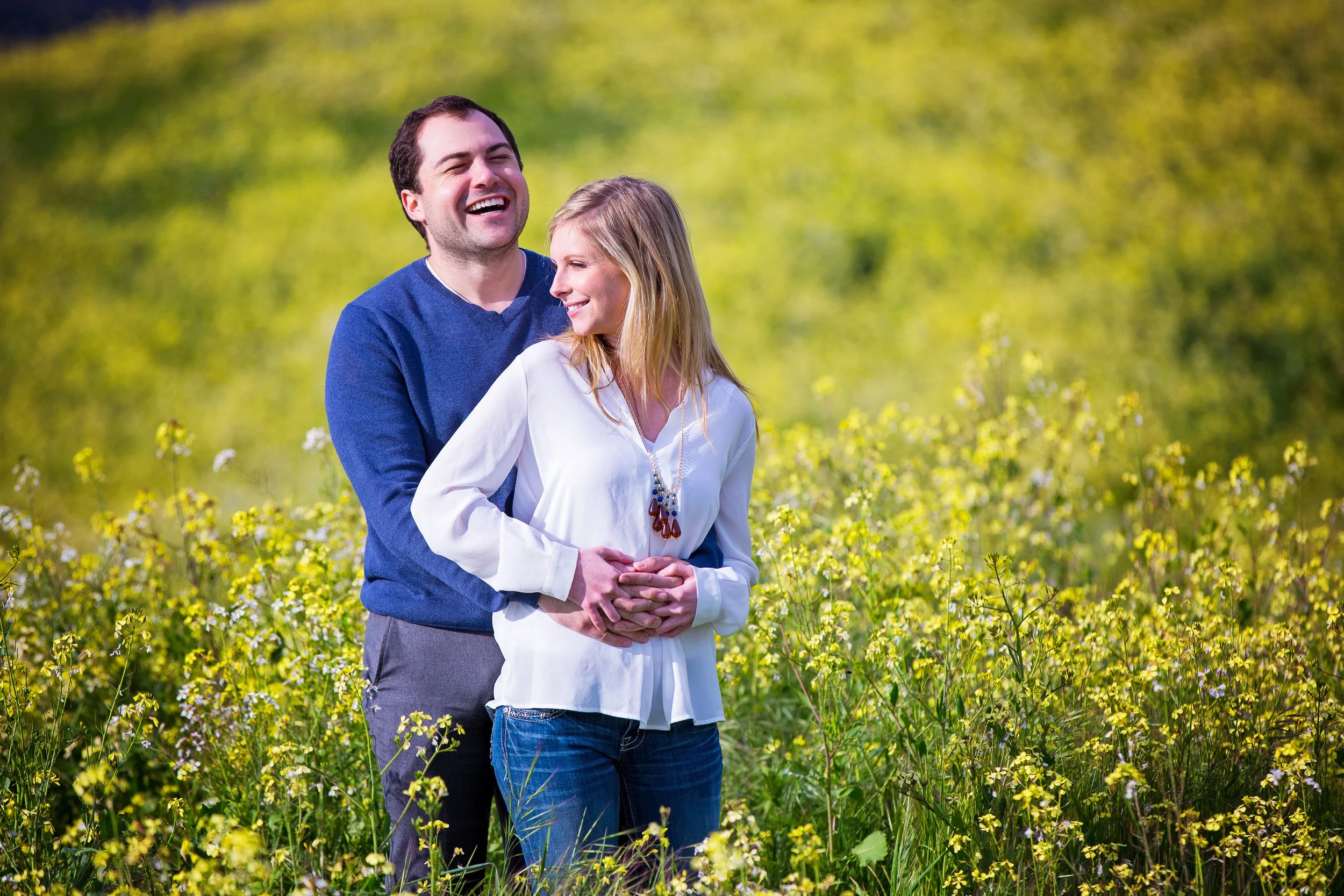 A smiling man in a blue sweater hugging a smiling woman in a white blouse with a necklace, standing in a field of yellow flowers with green trees in the background.