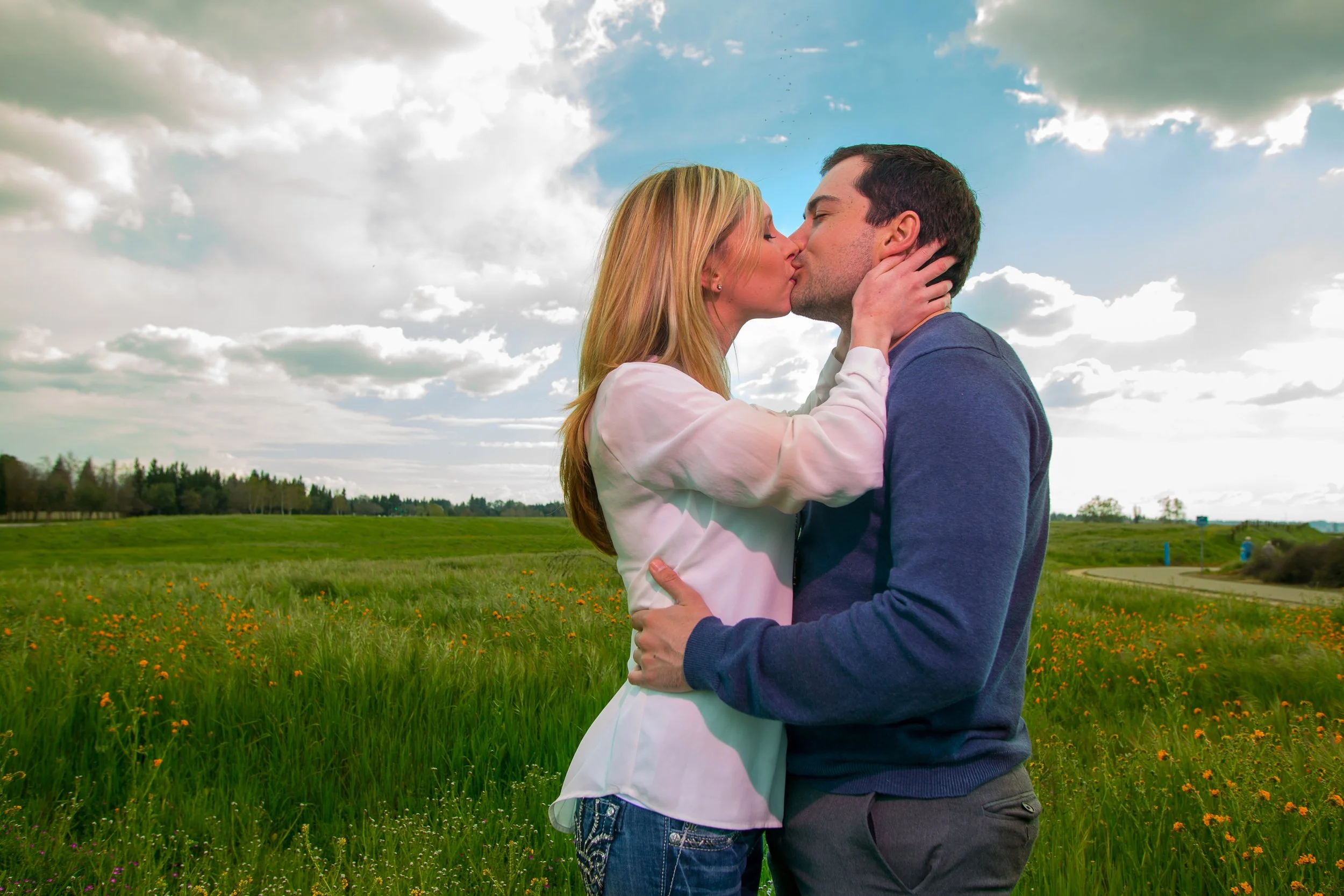 A couple kissing in a field with green grass and orange flowers, under a partly cloudy sky.