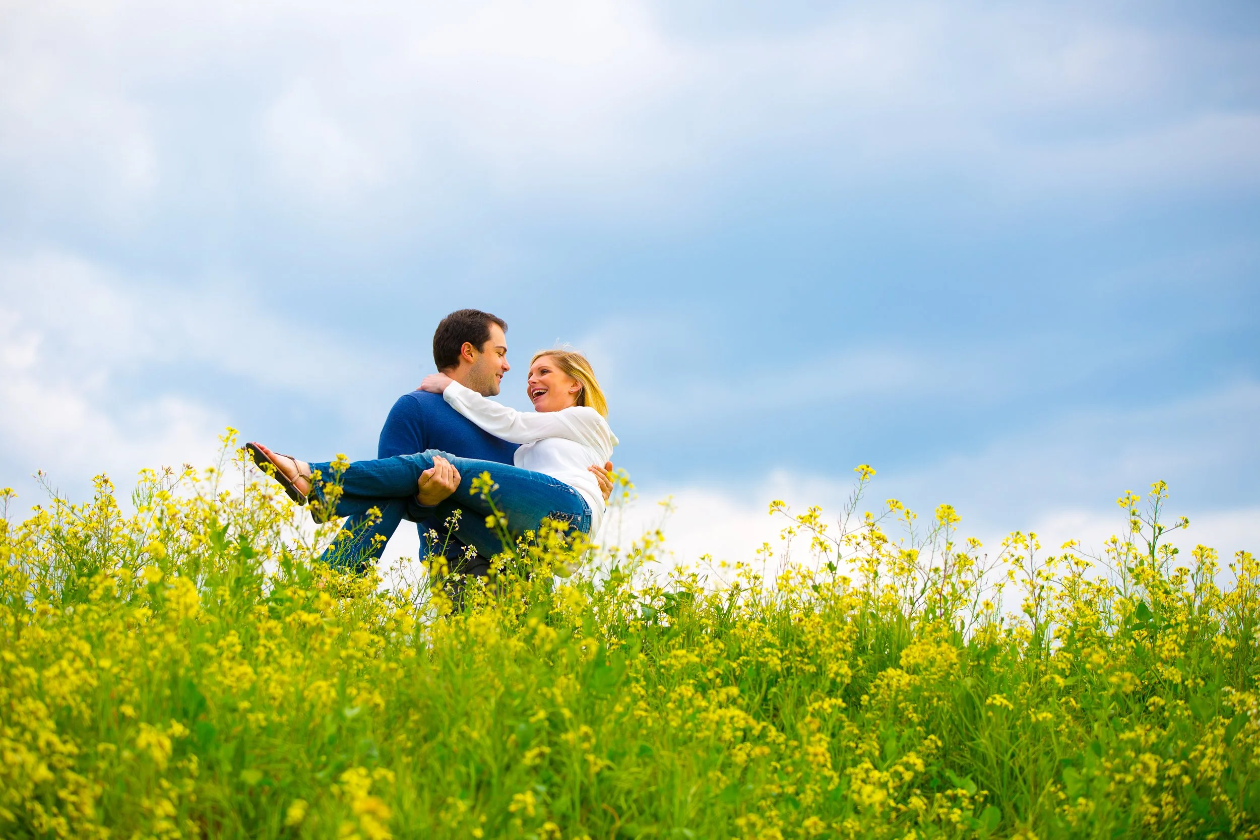 A couple in a field of yellow flowers, with the man carrying the woman in his arms. They are smiling and looking at each other against a blue sky with clouds.