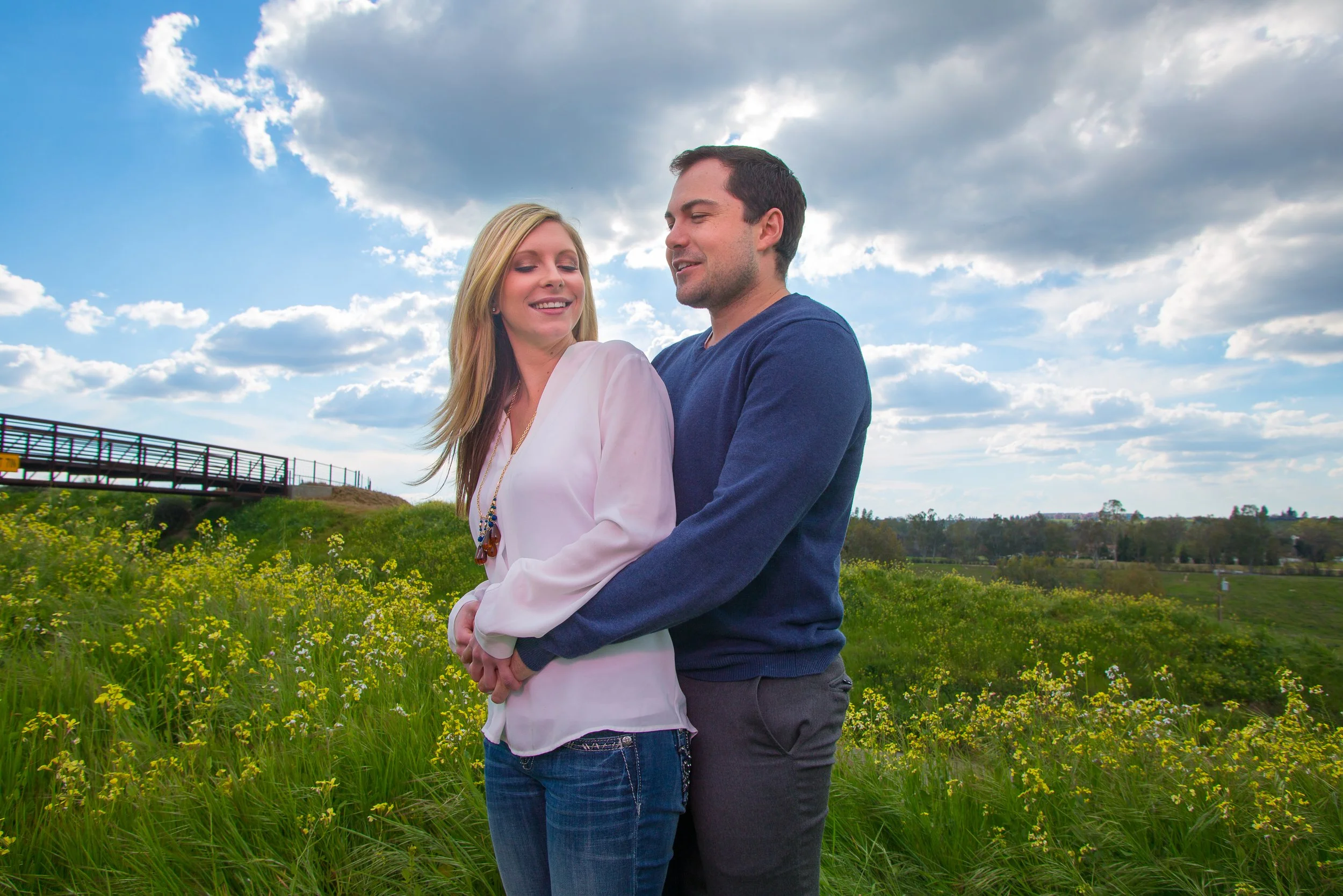 A happy couple standing together outdoors in a grassy field with yellow flowers, under a partly cloudy sky.