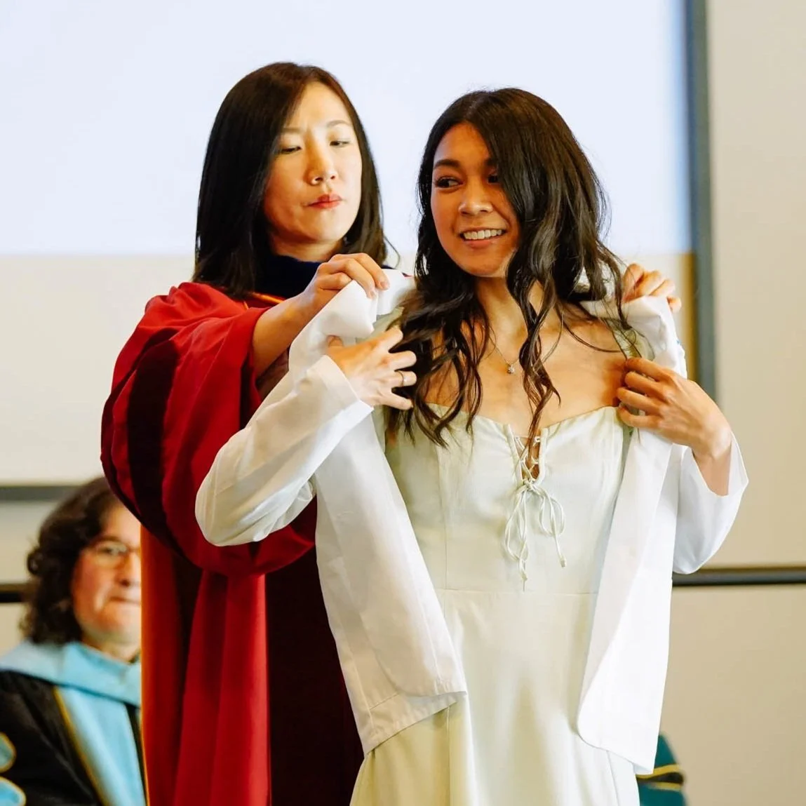 A woman in a white dress is being helped to put on a white jacket by another woman in a red gown. The woman in white is smiling while the woman in red is focused on helping her.