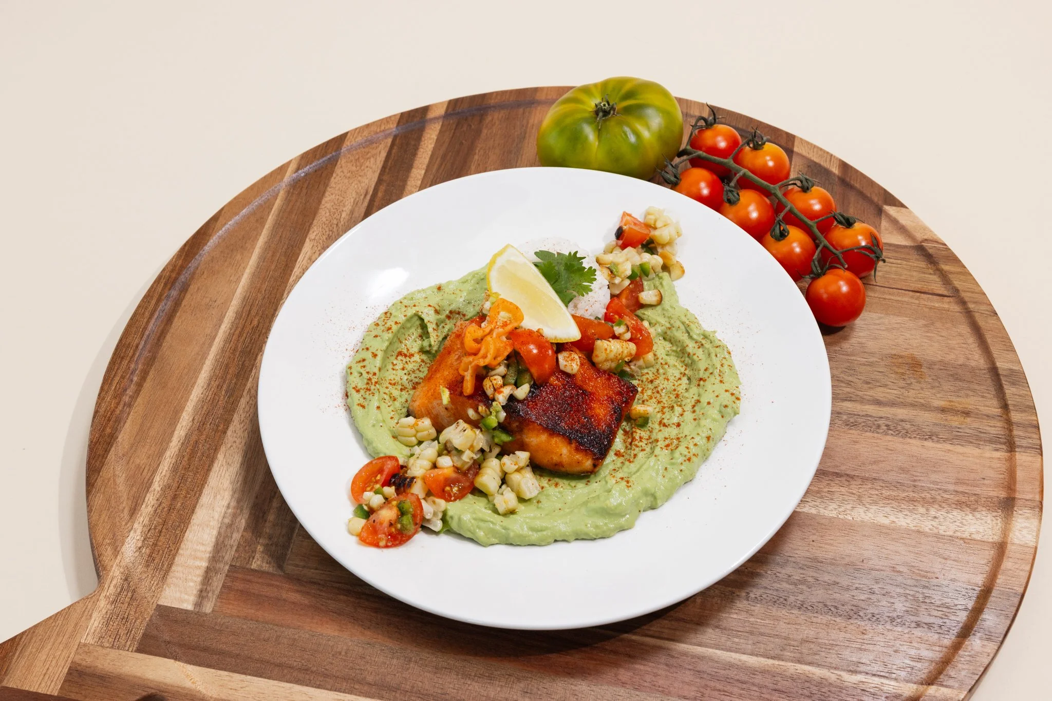 A white plate with grilled fish and rice with vegetables, garnished with lemon and herbs, on a wooden serving board with cherry tomatoes and a green heirloom tomato in the background.
