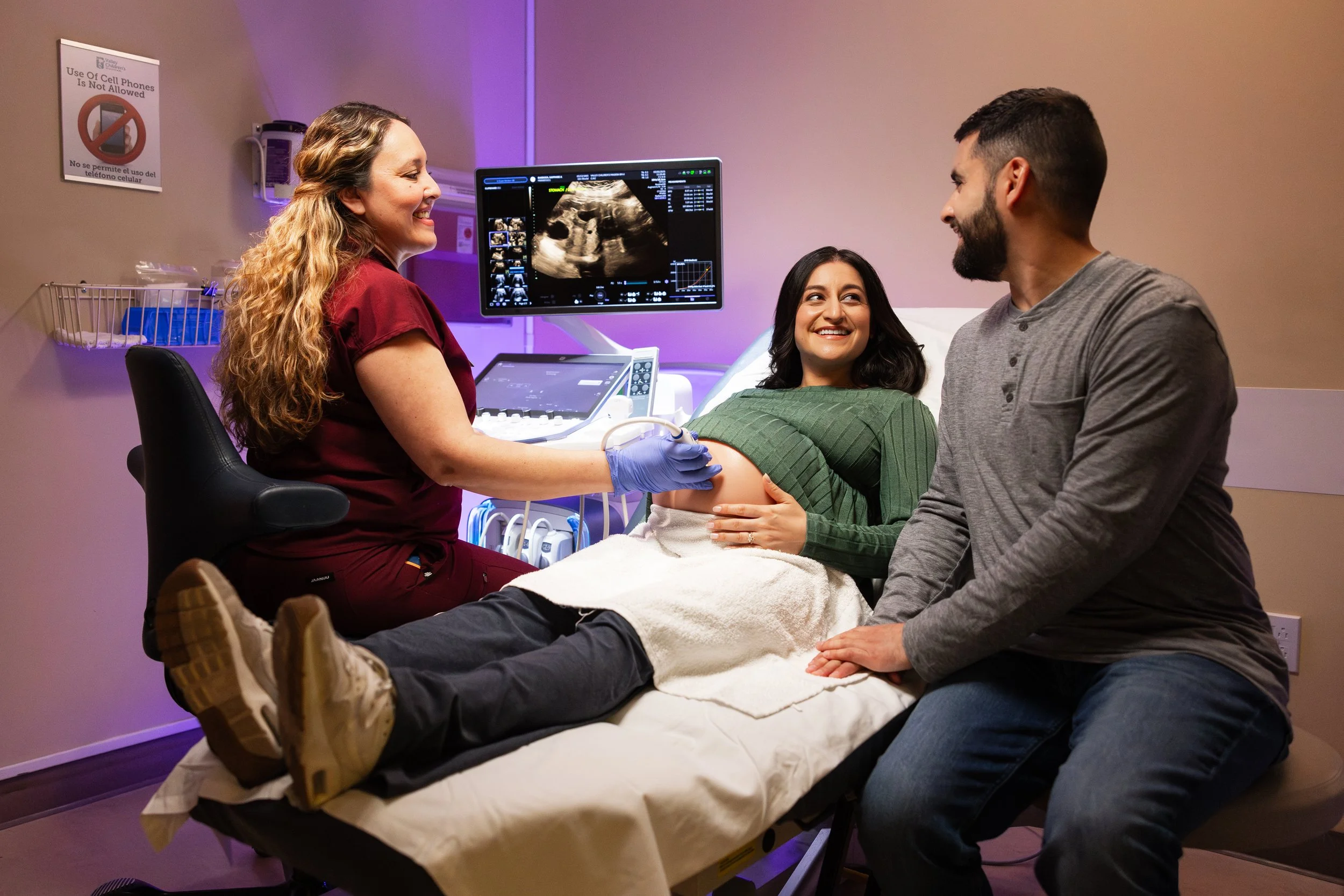 A pregnant woman lying on an examination table during an ultrasound appointment, smiling and holding her belly, with her partner sitting next to her. A healthcare professional is performing the ultrasound, which is displayed on a monitor behind them.