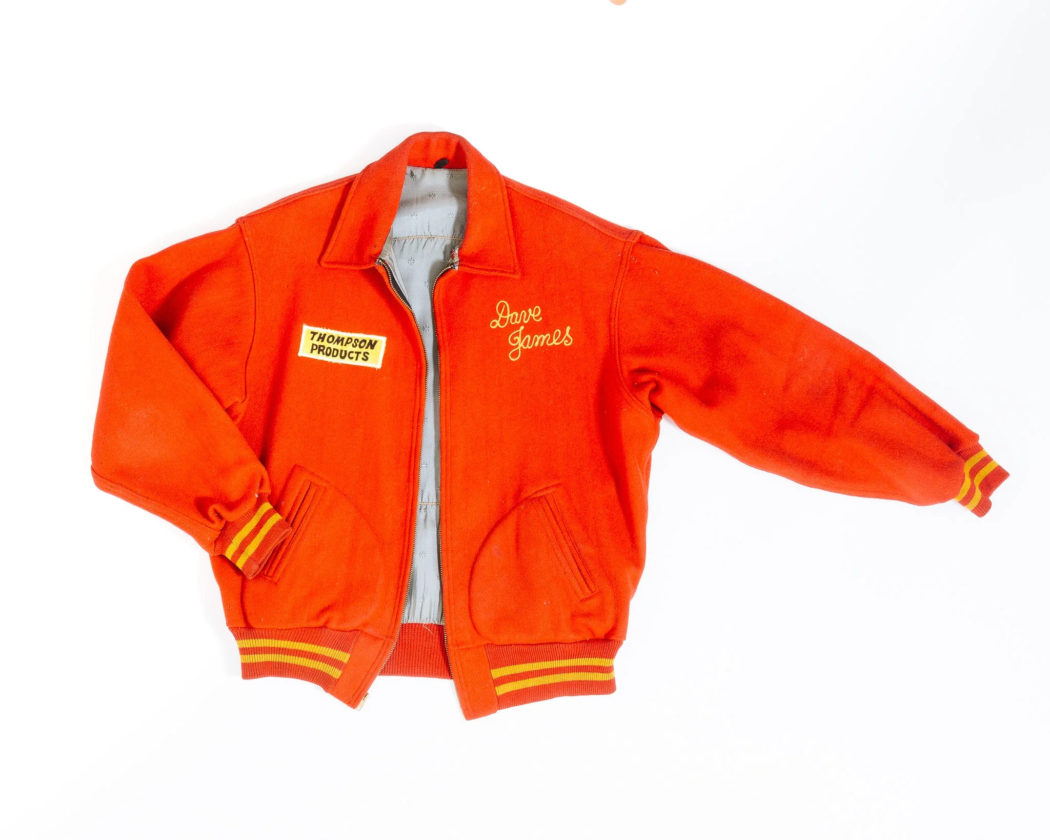 Red varsity jacket with yellow stripes on cuffs and a zipper in the front, embroidered with names and patches, laid flat on a white background.