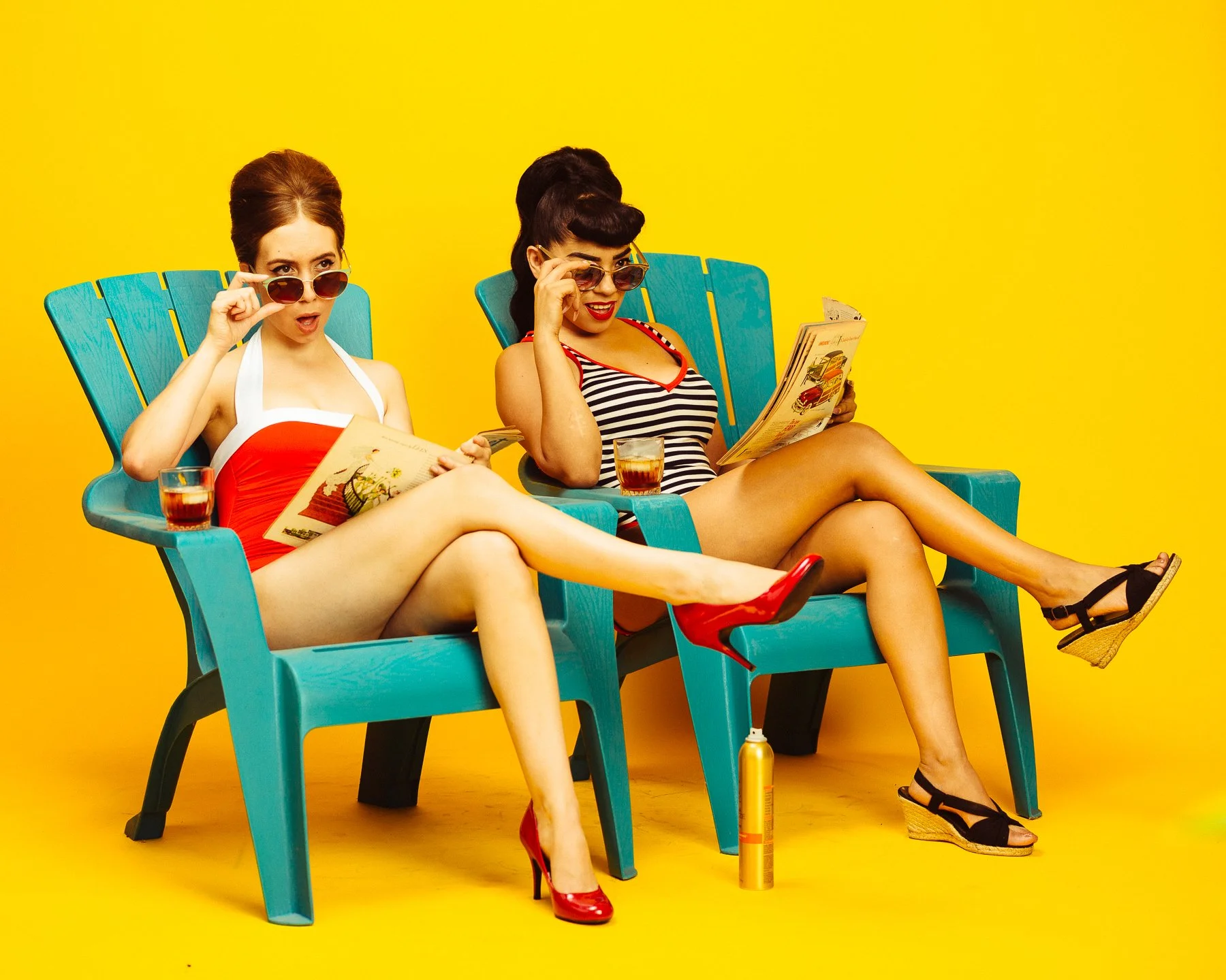 Two women sitting on blue lounge chairs against a yellow background, reading magazines and wearing sunglasses. One woman has red high heels, and the other has black wedge sandals with yellow soles.