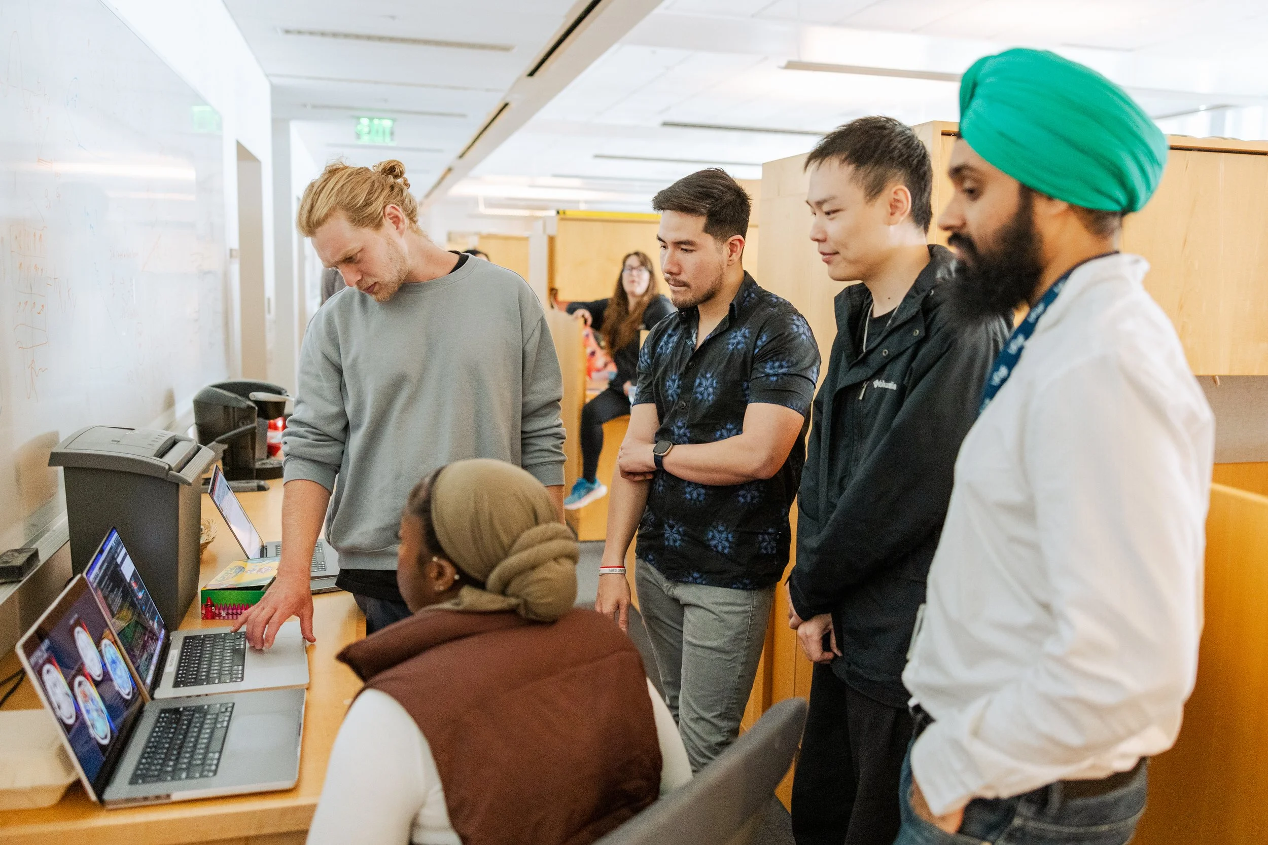 A group of diverse people standing in a line at a computer station in a modern office or classroom, with laptops and a whiteboard in the background.