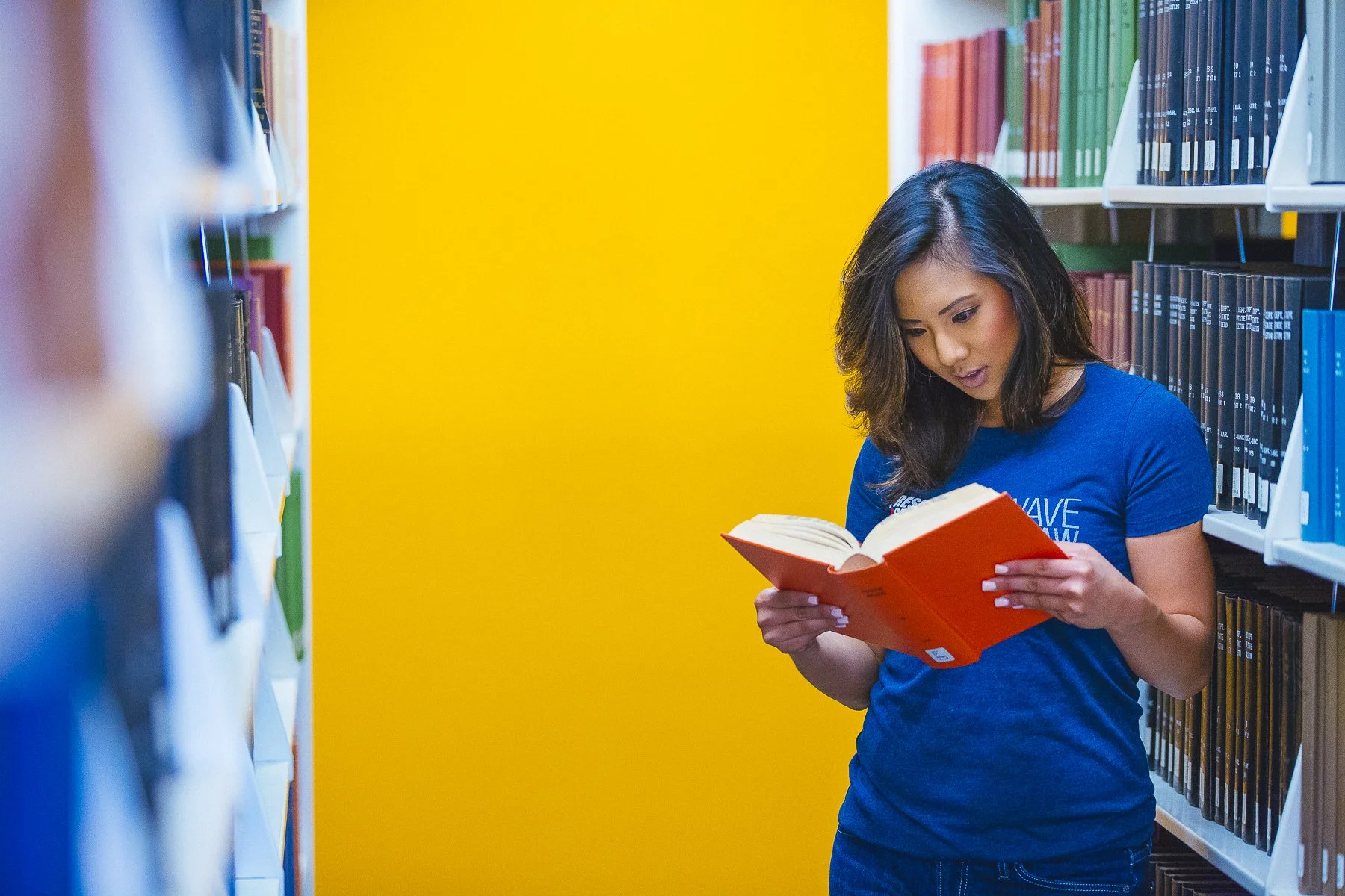 A woman with dark hair reading a book in a library, standing between yellow and white walls, surrounded by shelves of books.