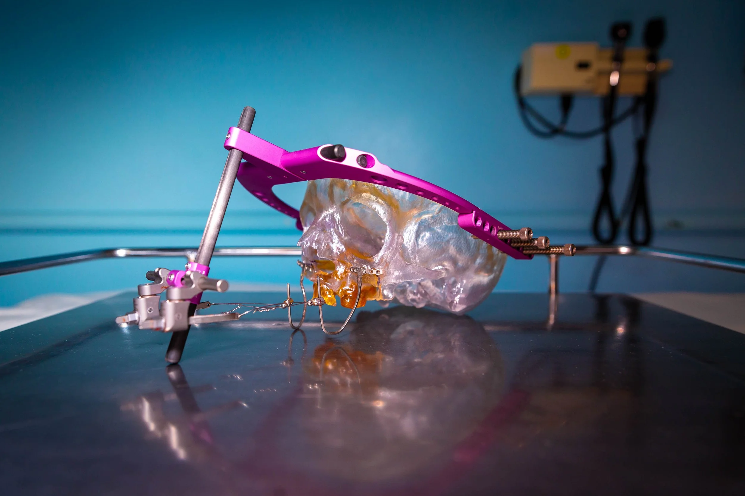 A surgical setup with a skull model secured in a pink surgical frame, positioned on a metal tray. In the background, there is a blurred medical device against a blue wall.