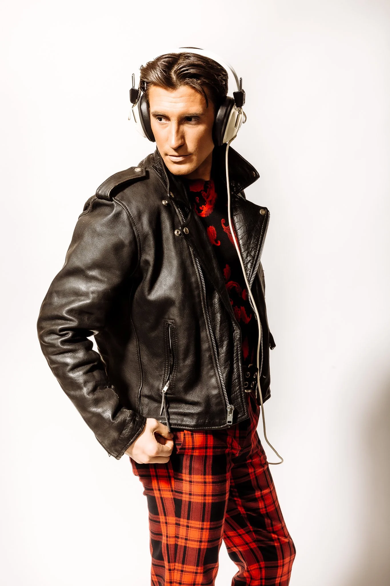 A young man with dark hair wearing a black leather jacket and red plaid pants, standing against a plain white background with headphones on.