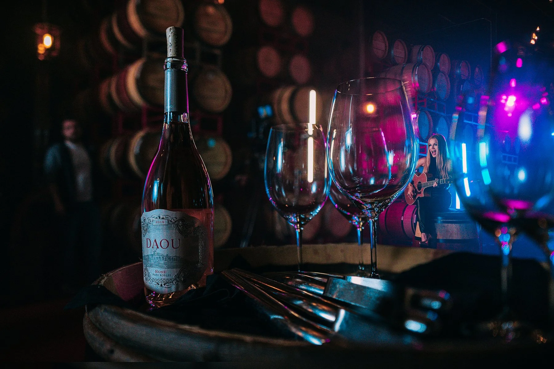 A bottle of red wine and three wine glasses on a table, with a live musical performance of a woman playing guitar in a dimly lit bar with wine barrels in the background.