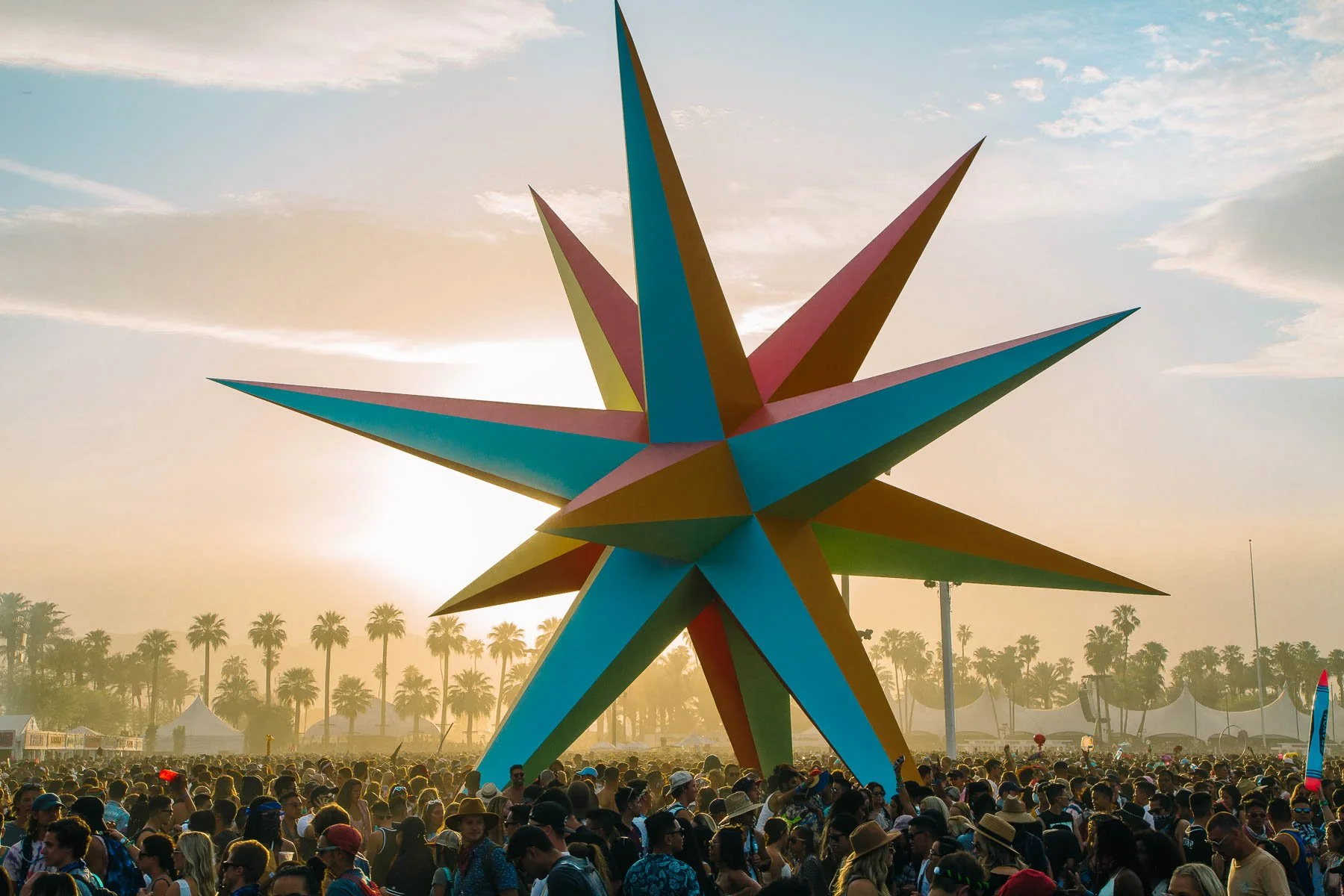 A large colorful star sculpture with multiple sharp points, set against a sunset sky during a large outdoor event with people and palm trees visible in the background.