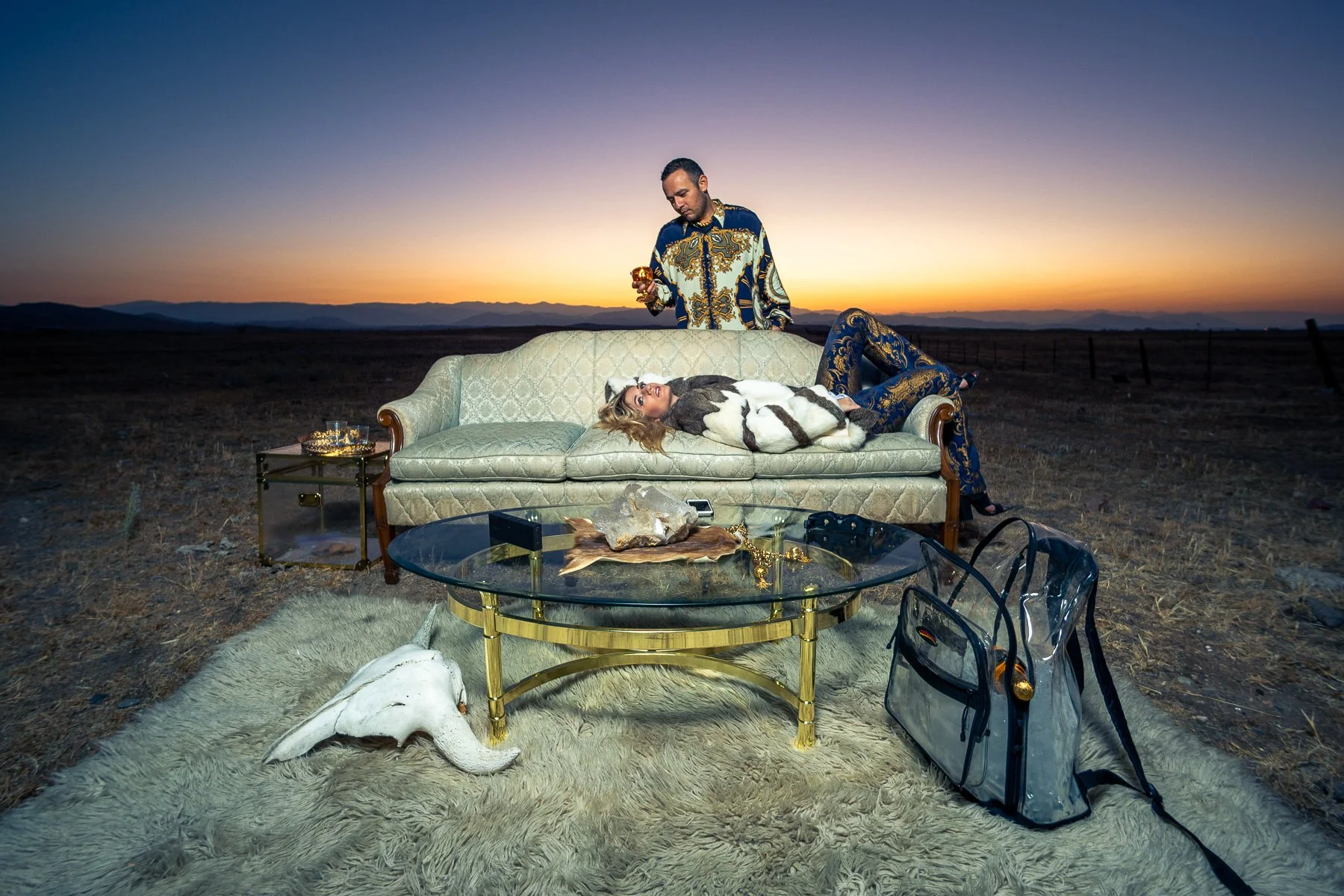 A woman lying on a vintage sofa in an open field at sunset, surrounded by luxury items like a glass coffee table, jewelry, and a skull, with a man standing nearby holding a candle.