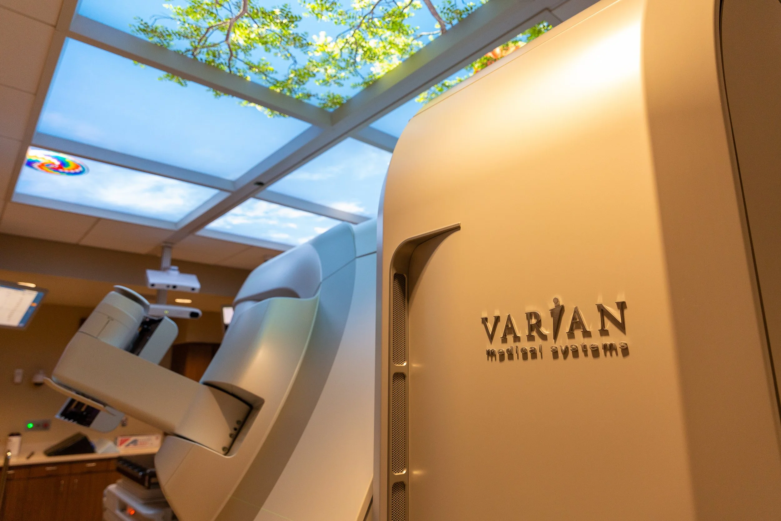 Interior of a medical facility with a large skylight showing trees and blue sky, featuring medical equipment and a sign for Varian Medical Systems.