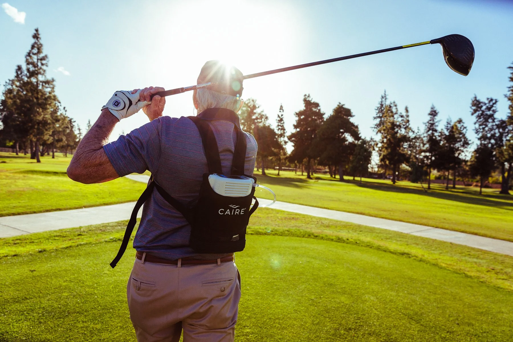 Older man playing golf on a sunny day carrying a golf club and wearing a golf glove, with trees and a sidewalk in the background.
