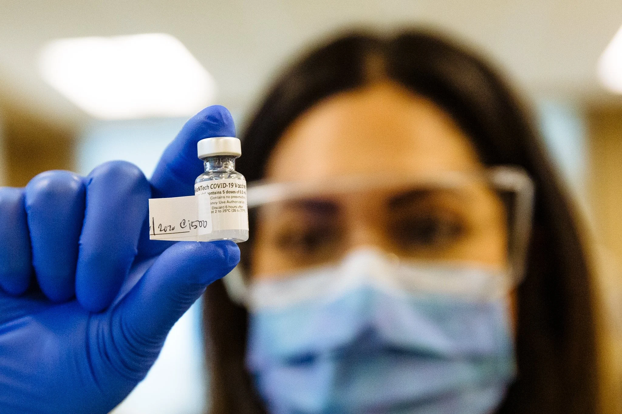 A person with dark hair wearing glasses and a face mask holds up a small vial labeled "COVID-19 Vaccine" in a clinical setting.