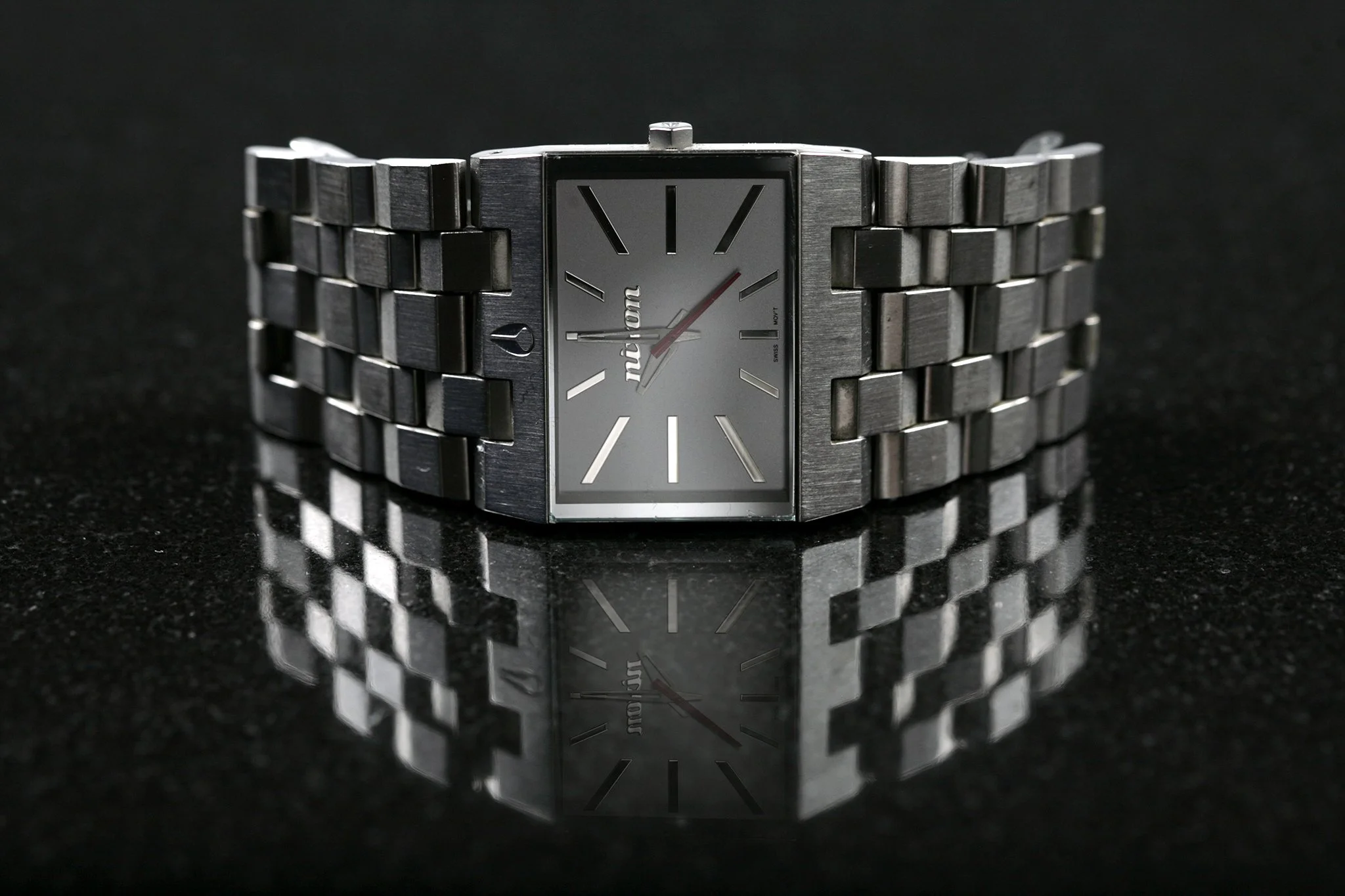 A silver rectangular wristwatch with a metallic band, black dial, and minimalist design, reflecting on a black surface with a textured background.