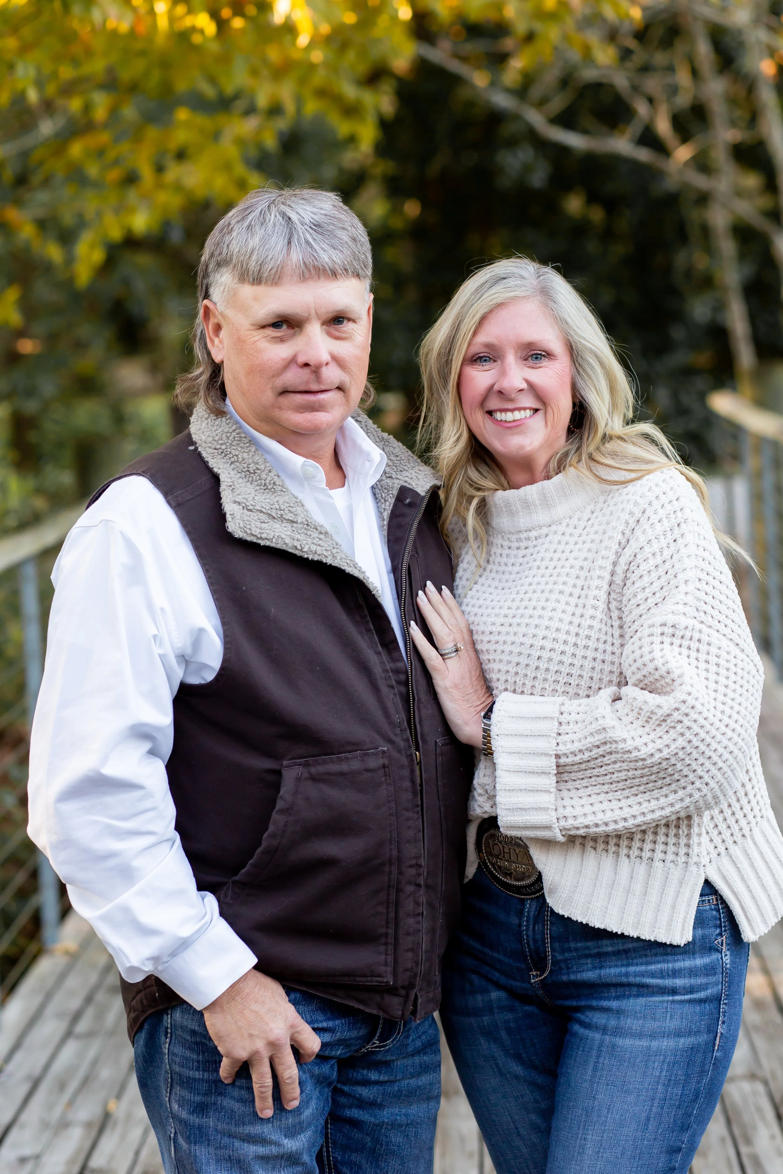A man and woman standing close together outdoors on a wooden walkway, with trees and yellow leaves in the background. The woman is smiling and has her hand on the man's chest.