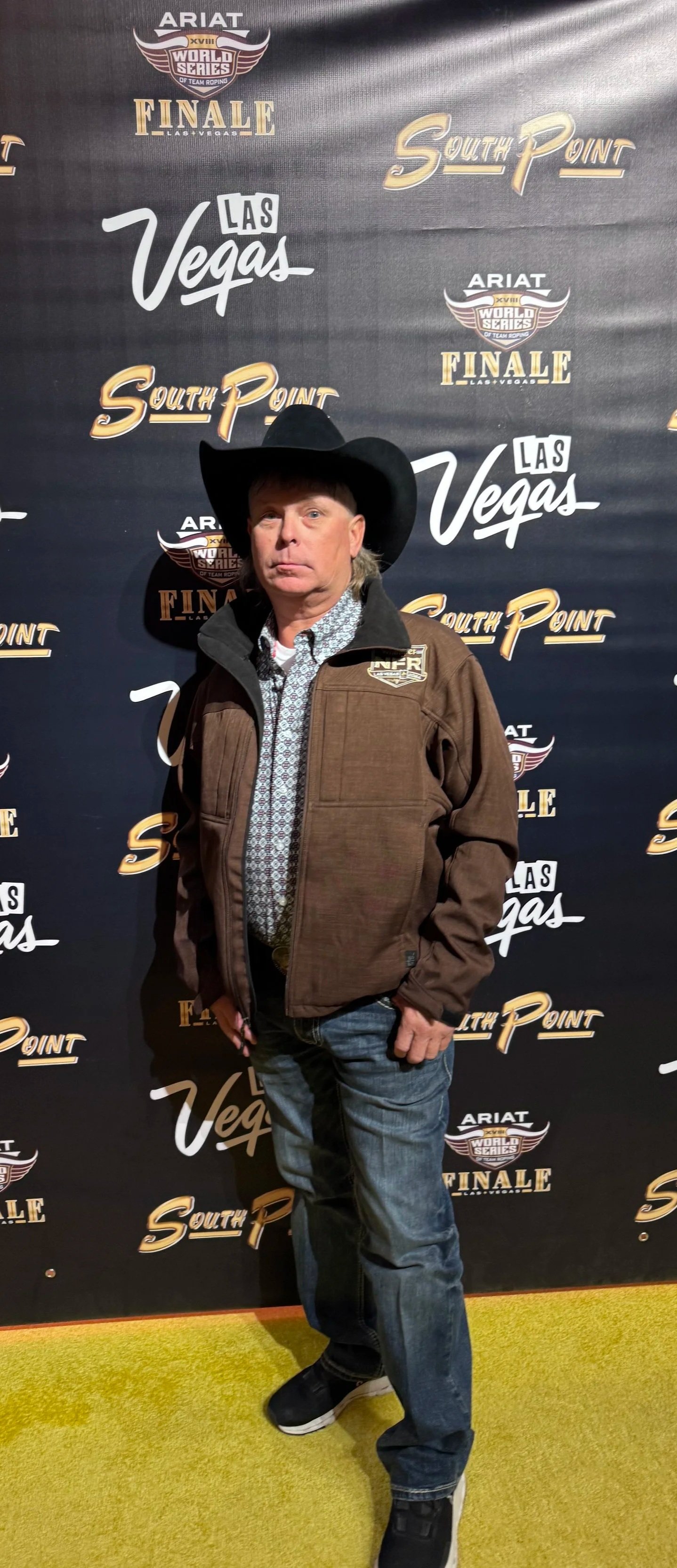 A man wearing a cowboy hat, brown jacket, patterned shirt, and jeans stands in front of a step-and-repeat banner with logos for the World Series of Team Roping, South Point, and Las Vegas.