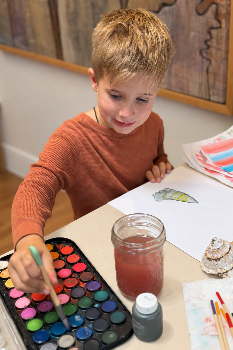 A young boy with blond hair and a brown shirt painting with watercolors at a table. There are watercolor paints, a jar of red water, a seashell, and paper with watercolor paintings on the table.