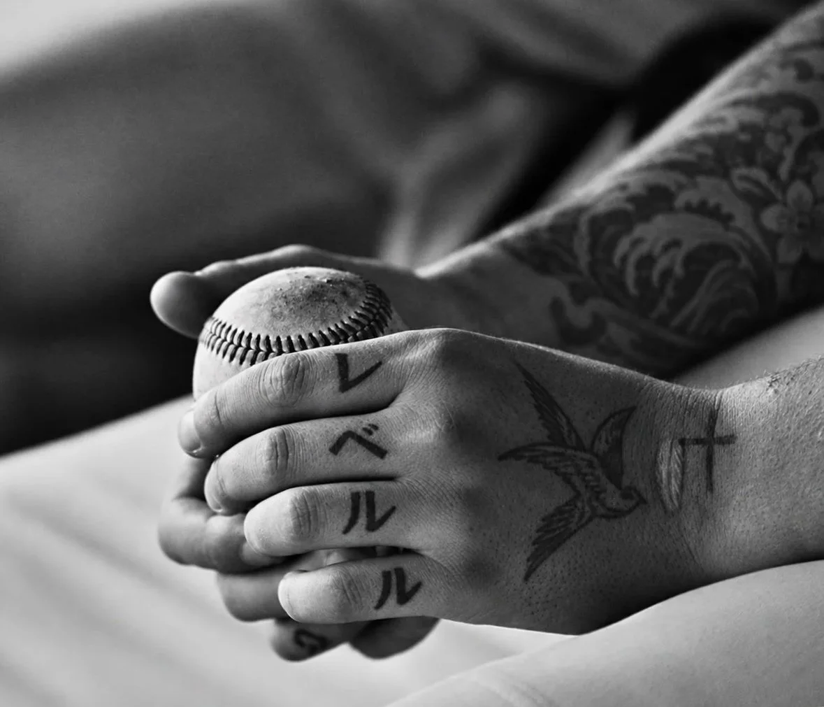 Close-up of a person's tattooed hand gripping an aged baseball, with additional tattoos visible on the person's arm and wrist.