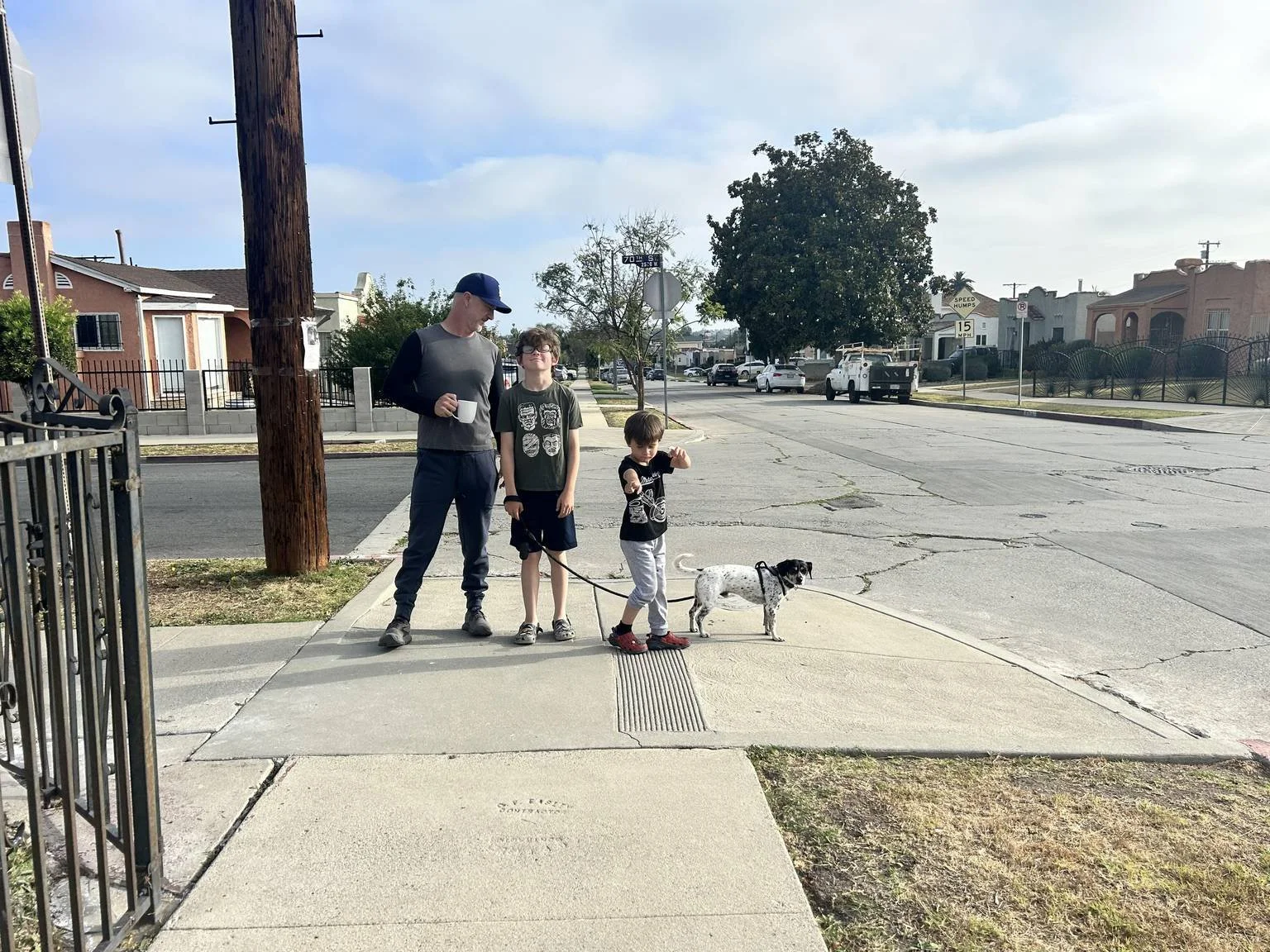 A man and two boys walking a small dog on a sidewalk in a neighborhood, with houses, parked cars, and a street sign in the background.