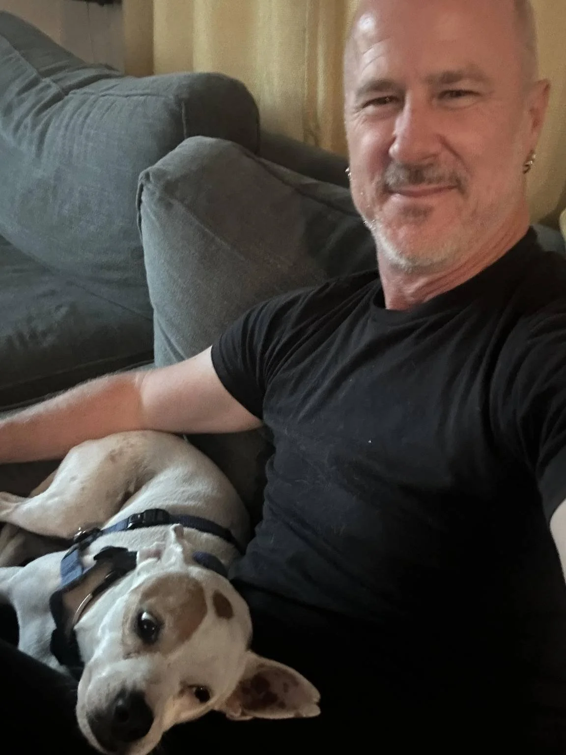 A man smiling while sitting on a couch with a Chihuahua dog resting on his lap.