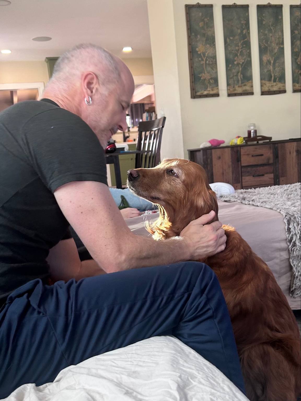 A man with short hair, earrings, wearing a black t-shirt, is sitting on a bed with a golden retriever dog. The man is smiling and holding the dog gently by the neck. The dog sits calmly, looking at the man. The background shows a living room with a sideboard, wall art, and some household items.