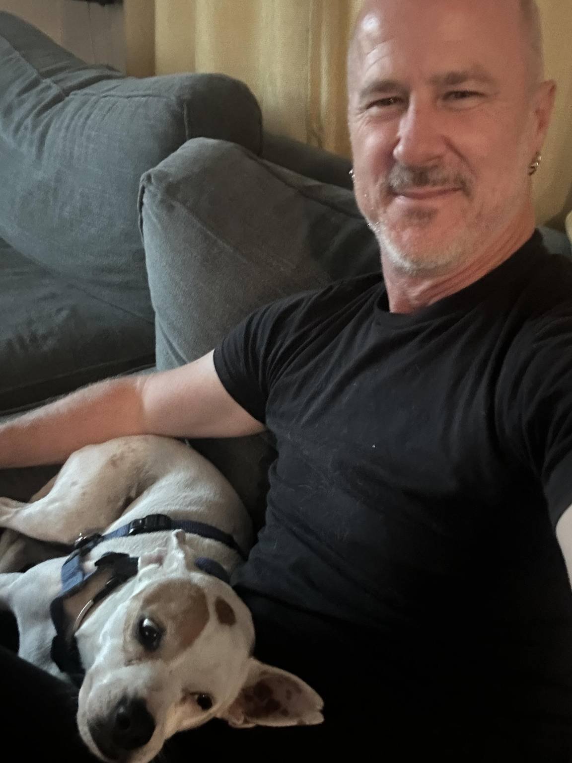 A man smiling with a dog lying next to him on a couch.