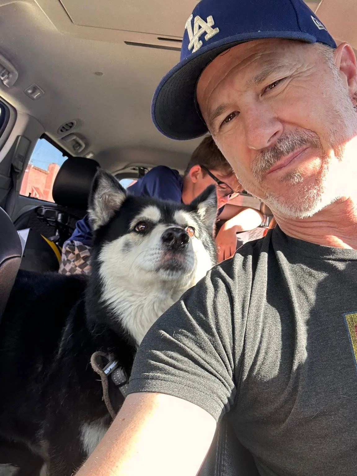 A man taking a selfie inside a vehicle with two other people, a husky dog, and a person in the background. The man is wearing a blue Los Angeles Dodgers cap and a dark T-shirt. The dog has black and white fur and looks at the camera.