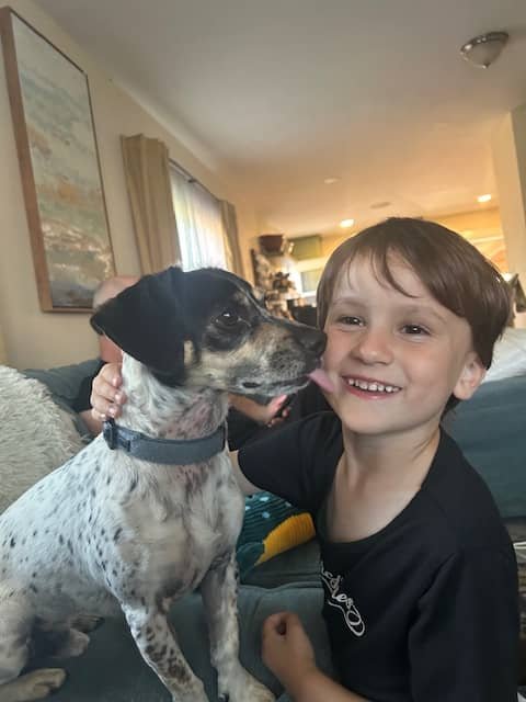 A young boy smiling as a dog licks his face inside a cozy living room.