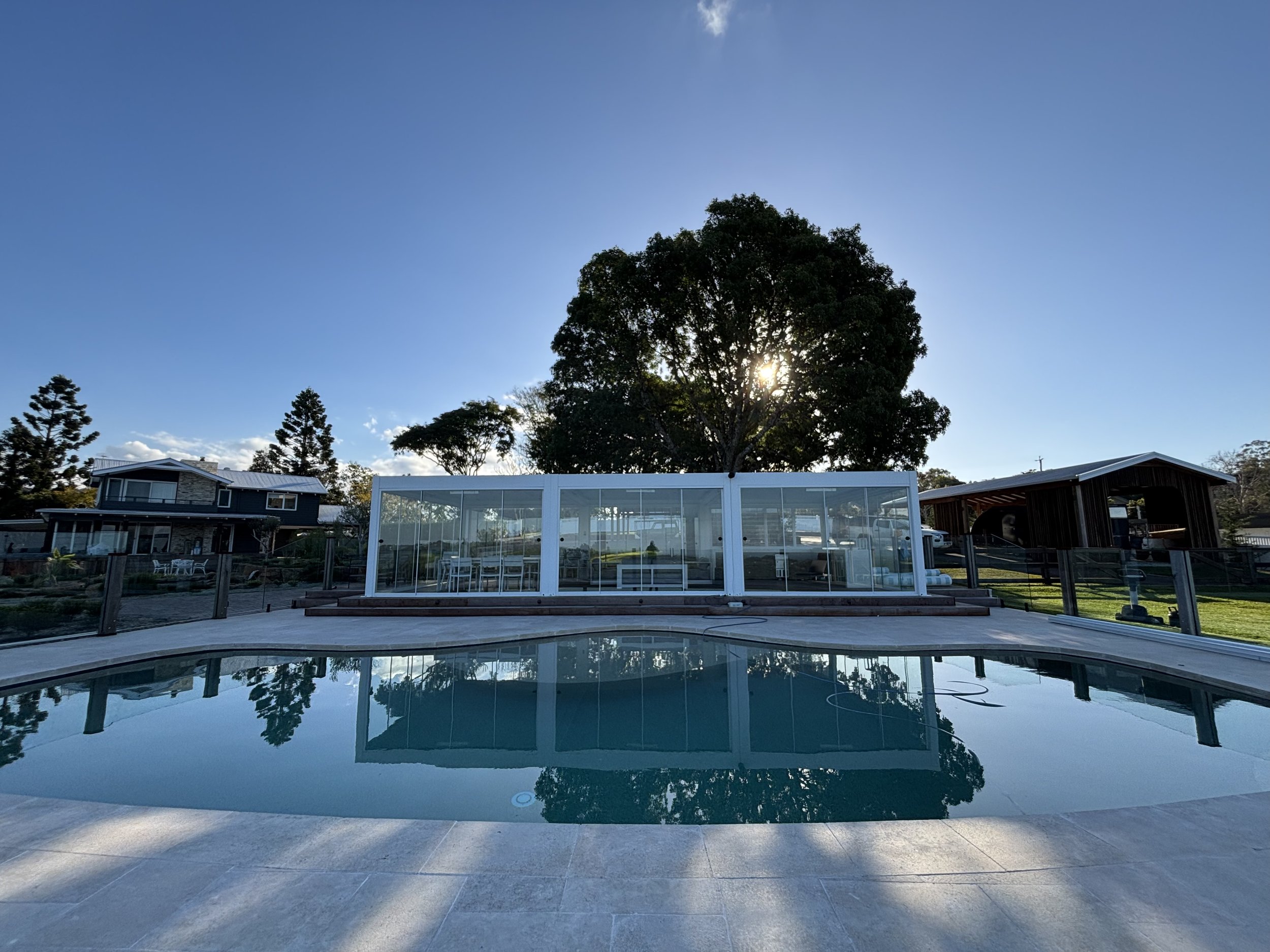 View of a backyard with a swimming pool in the foreground, surrounded by a patio, with a house, trees, and a clear blue sky in the background. The sun is peeking through a large tree.